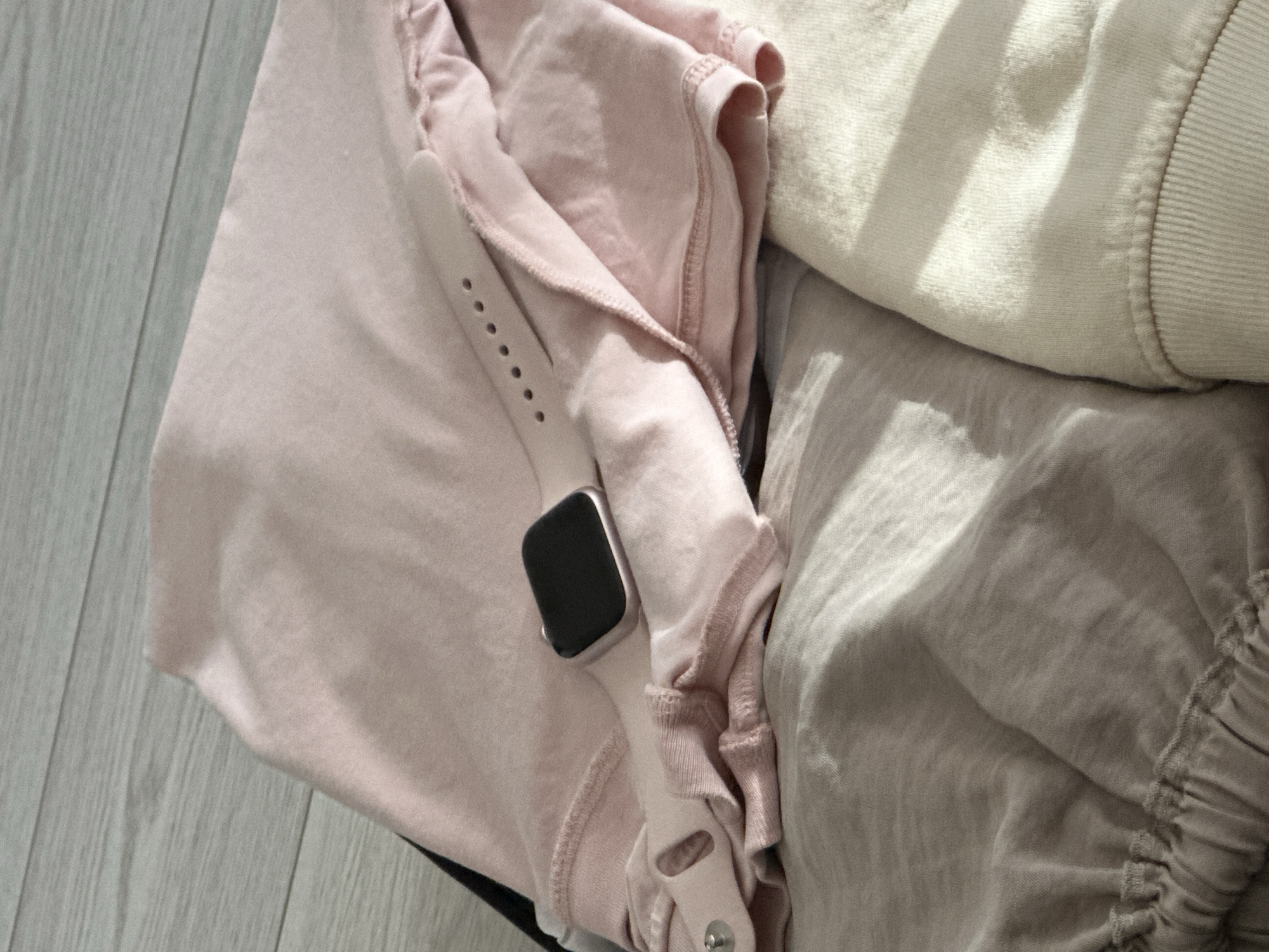 The image shows a close-up view of several folded clothing items and a smartwatch. The items appear to be a pink t-shirt or top, a light grey pair of pants or skirt with an elastic waistband, and a cream-colored sweatshirt or hoodie. The smartwatch has a light pink band and a dark rectangular face, and is resting on the pink top. The clothing is laid out on a light-colored wooden floor. There are no people or any specific indications of a location within the city of Sofia, Bulgaria.