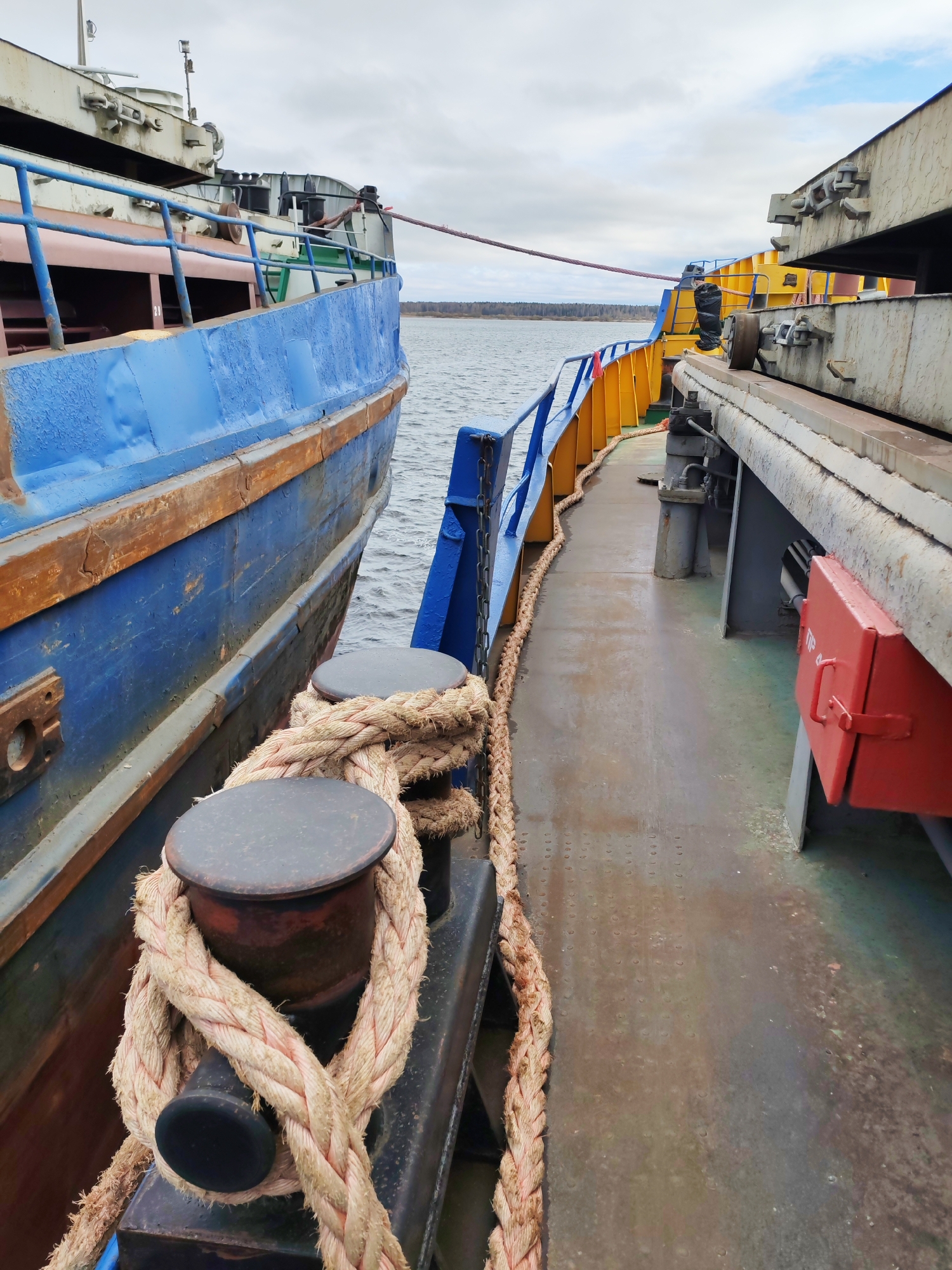 The image shows a close-up view of two boats docked side-by-side. A thick rope is wound around two bollards on the boat in the foreground. The boat on the left is painted blue with visible rust. The boat on the right has a yellow railing and a red storage box on the side. In the background, a body of water can be seen with trees lining the opposite shore. The sky is overcast. Based on the composition and visible characteristics, the image might be taken in Rybinsk, Russia.