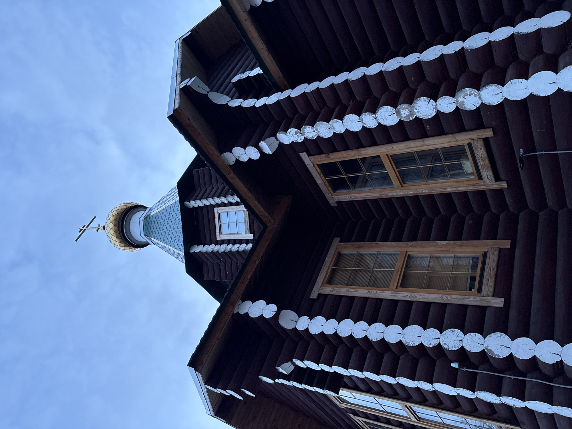 The image captures an upward-looking view of a traditional wooden building, likely an Orthodox chapel or church, set against a bright, partly cloudy blue sky. The building is constructed from dark brown logs, giving it a rustic aesthetic.
A prominent architectural feature consists of decorative rows of white-painted, round log cross-sections that adorn the eaves and cornices, creating a distinctive scalloped pattern. Multiple rectangular windows with light-colored wooden frames and grid-like panes are visible within the dark log walls.
Towards the upper part of the structure, a golden, onion-shaped dome (cupola) with a prominent three-barred Orthodox cross on top stands out. Below the golden cap, the dome features a patterned, possibly tiled or shingled, surface in a muted green hue.
The scene depicts the building's exterior during daytime, with clear, soft lighting. There are no people or specific activities taking place. The architectural style, characterized by log construction and an Orthodox dome and cross, is consistent with the provided location context of Chaikovsky, Russia. No visible text is present in the image.