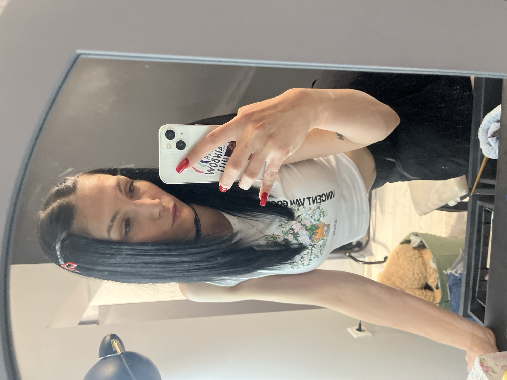 A young woman with long, dark hair is captured in a mirror selfie, holding a white smartphone with red polished nails. She wears a white t-shirt featuring a reversed graphic that appears to say "VINCENT VAN GOO" (likely "Vincent van Gogh") with a floral design, and black bottoms. A black choker necklace is visible around her neck, and a small red hair clip adorns her hair. The smartphone case shows a reversed colorful "RAINBOW" design. A small tattoo is visible on her right arm. The indoor setting features a dark gray wall reflected in the mirror's frame, with a black lamp visible in the lower left. In the background, partially obscured, are a light brown fluffy object (possibly a dog or a blanket) and a light blue rolled-up towel or fabric. The woman has a neutral expression, looking into the camera.