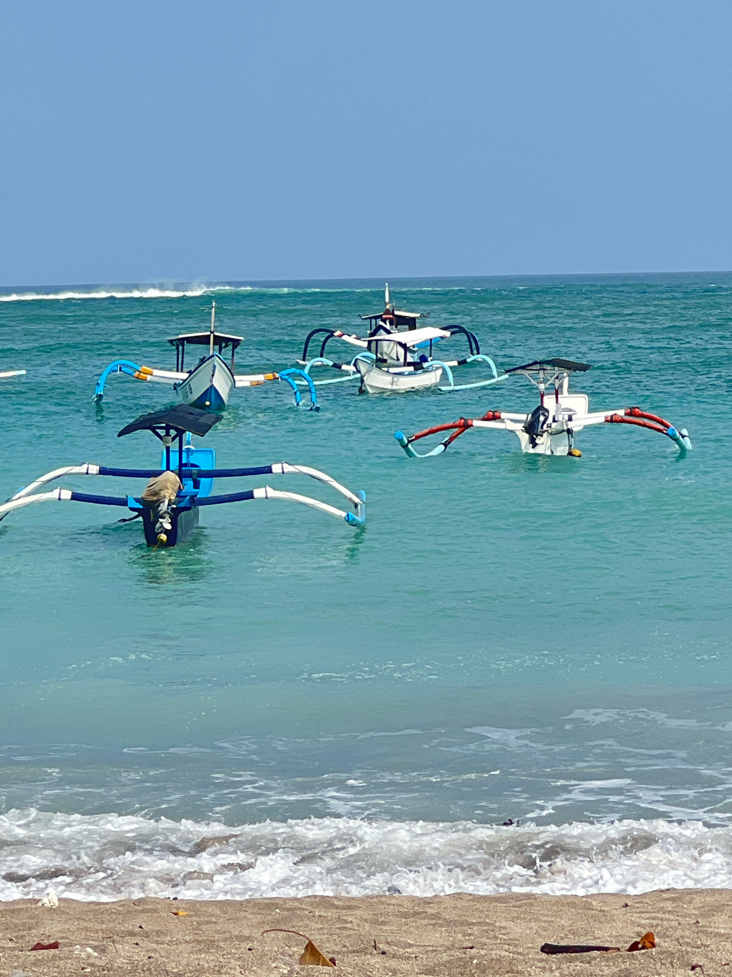 A serene scene unfolds in Kuta, Indonesia, where the turquoise waters of the ocean meet a sandy shore. Several traditional outrigger boats, known as jukungs, are anchored offshore. The boats, with their distinctive stabilizing outriggers, are a common sight in this region. One jukung in the foreground has a tan bag draped over its motor. The water is calm, with gentle waves lapping at the beach. The sky is a clear, brilliant blue, indicating a bright and sunny day. The overall impression is one of tranquility and natural beauty.