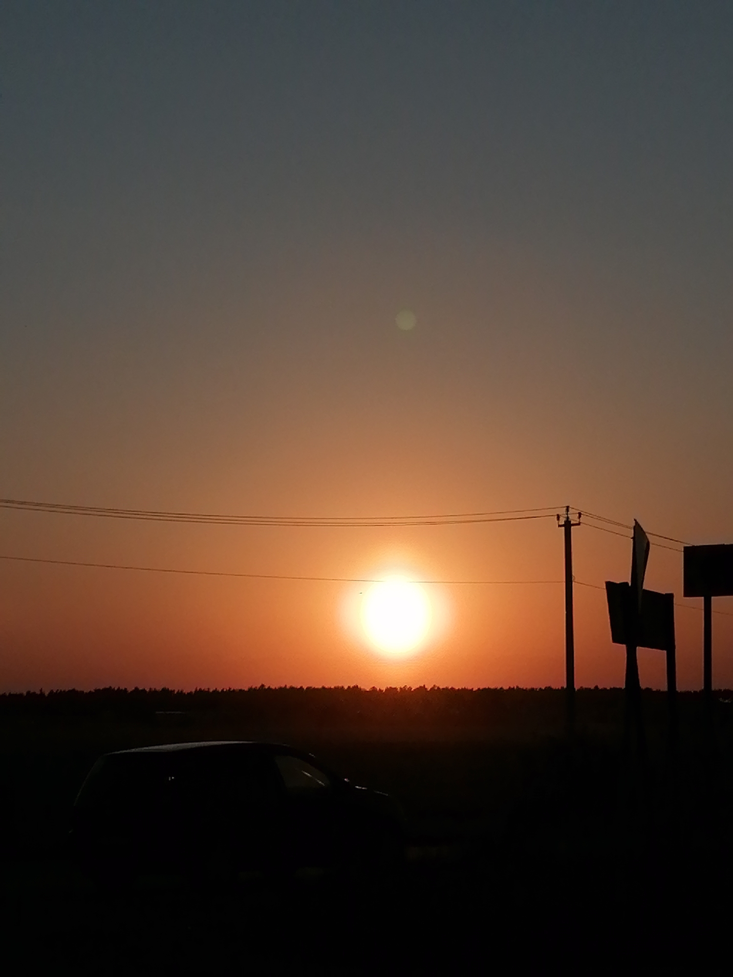 The image depicts a sunset in an unidentified city, with a low horizon line obscured by dark vegetation and silhouettes of utility poles and road signs. A vehicle is parked in the foreground, its shape barely discernible against the dim foreground. The sky transitions from a dark blue at the top to a vibrant orange near the sun, which is positioned centrally and casting a bright glow. Overhead wires stretch across the horizon.