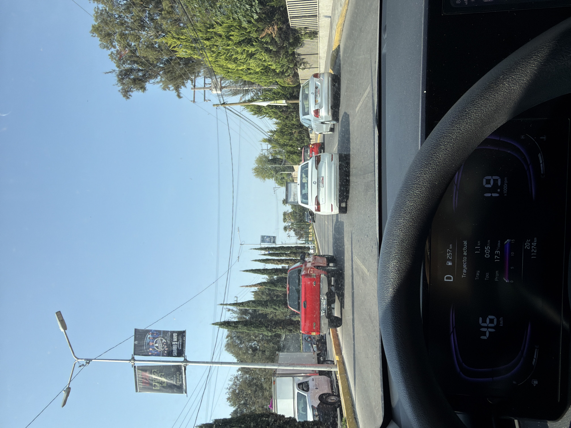The image, likely taken from inside a car in Puebla, Mexico, shows moderate traffic on a paved street lined with trees and buildings. Several vehicles, including a red pickup truck, white trucks, and silver sedans, are visible in the traffic ahead. A lamppost with banners, possibly advertising an event, stands on the left side of the street. The car's dashboard is partially visible, displaying a digital speedometer reading 46 km/h and other trip information. The weather appears sunny, indicated by the clear blue sky.