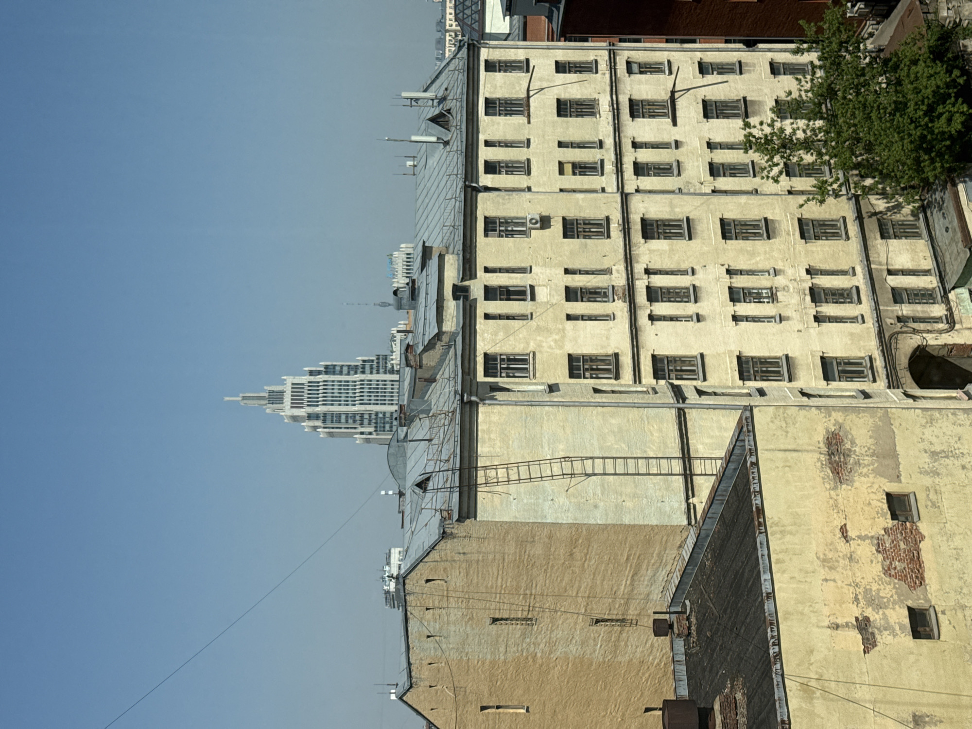 This image presents a cityscape, featuring a view of several buildings against a clear, blue sky. The most prominent building is a large, older, multi-story structure with rows of windows that appear to be boarded up. The building is painted in a faded yellow, showing signs of aging and weathering. A smaller building with similar weathered paint and design is situated in front of it. On top of the smaller structure is a rusty roof. A ladder connecting the roofs of adjacent buildings can also be seen. In the background, a modern skyscraper with a distinct spire stands out. Based on the architectural styles of the buildings, particularly the historical building with many boarded up windows, the modern skyscraper in the background, the photo most likely depicts a location in Moscow, Russia.