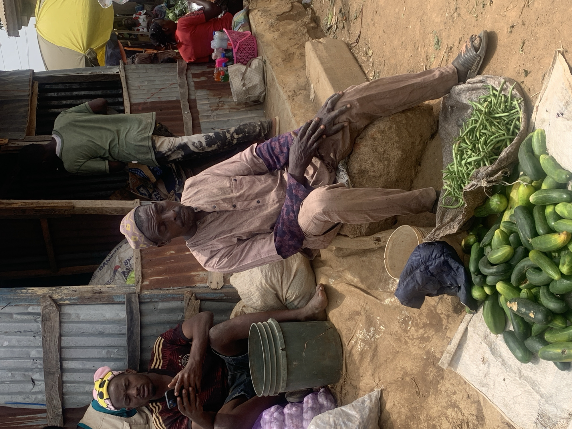 The image shows a market scene in Jos, Nigeria. Two men are visible in the foreground, sitting on the ground. One man is leaning back against a pile of vegetables, including cucumbers and green beans. He is wearing a light pink shirt, dark trousers, and a patterned cap. His feet are propped up on a rock and a sack of green beans. Next to him, another man is sitting on the ground, wearing a dark shirt, shorts, and a hat. A bucket rests near him. To the upper left, another man is visible standing inside of a simple, tin-roofed building, wearing a green shirt and pants. There are some people above the roof wearing red and yellow. The ground is bare dirt. A white sheet of fabric is also seen.