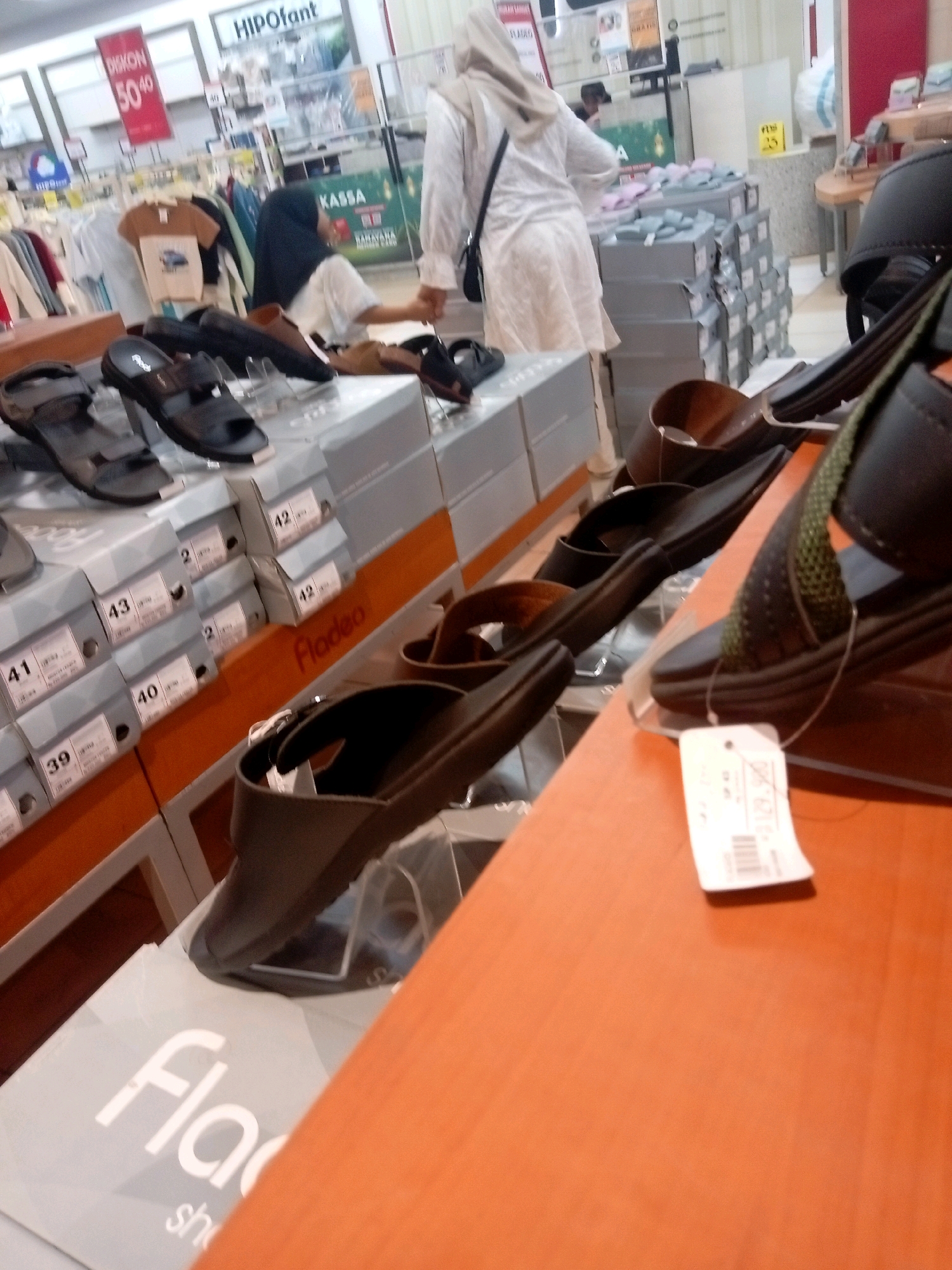 The image depicts a retail store environment, likely a shoe store, in Batam, Indonesia. The scene is filled with display shelves stacked with shoe boxes and shoes, primarily sandals and flip-flops. A woman wearing a hijab and an off-white dress is visible in the background, seemingly browsing or interacting with the merchandise. Several stacks of gray shoe boxes are prominent, with some bearing labels that indicate sizes like "41," "40," and "39." On the foreground shelf, there are multiple pairs of dark-colored sandals on display, some with price tags attached. One price tag clearly shows a price of "Rp 129.900". The overall lighting suggests an indoor, well-lit commercial space. The store appears to be part of a larger establishment, with signage like "HIPOfant" visible in the background.