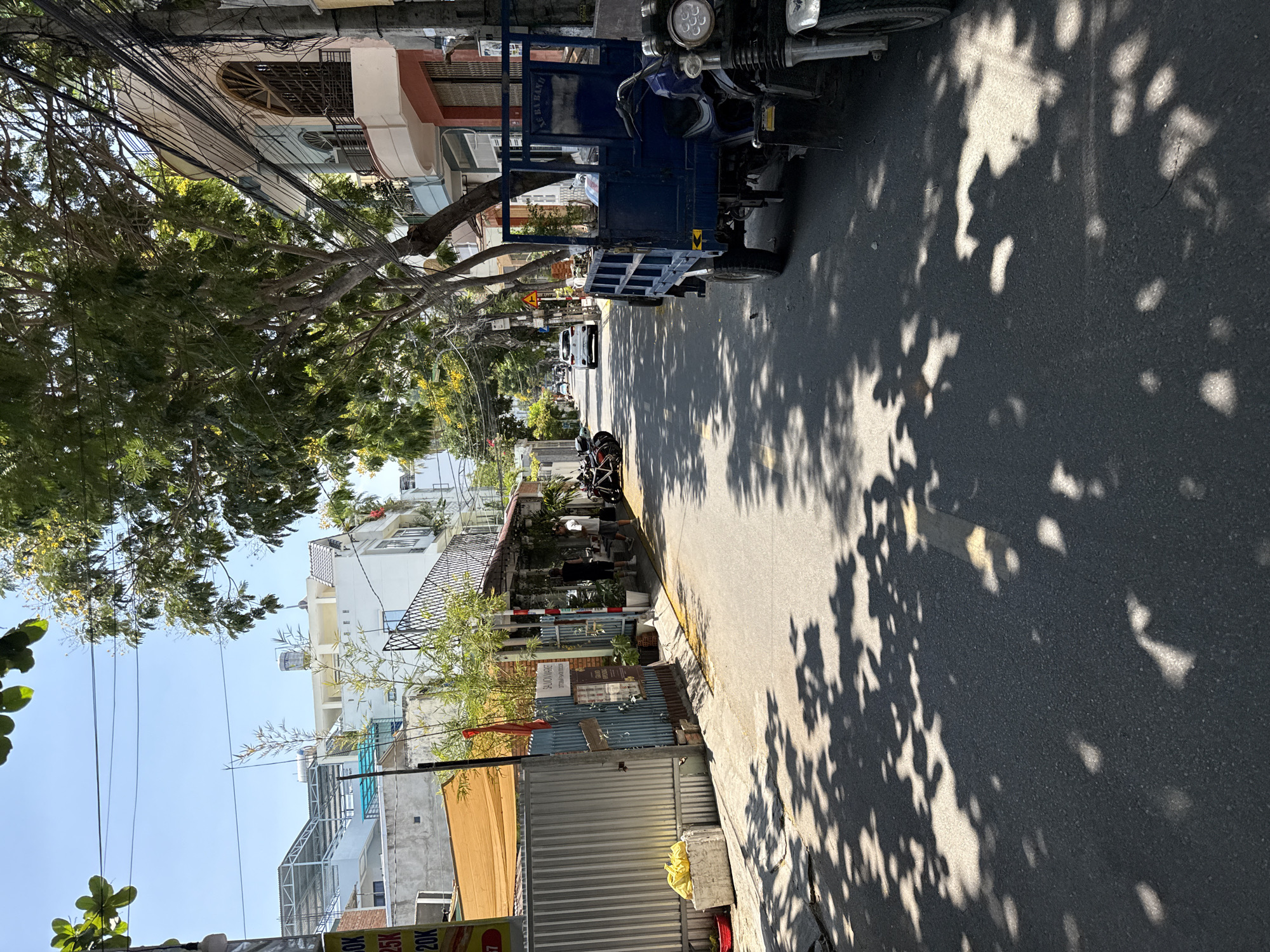 The image depicts a street scene in Bắc Nha Trang, Vietnam. The view is taken from the street level looking down the road. Buildings line both sides of the street. Wires are strung across the street. A tree overhangs the street on the left, casting dappled shadows on the asphalt. A blue truck is parked on the right side of the street. Several motorbikes are parked further down the street. People are visible on the sidewalk in front of a business on the right. A white car is visible further down the street.