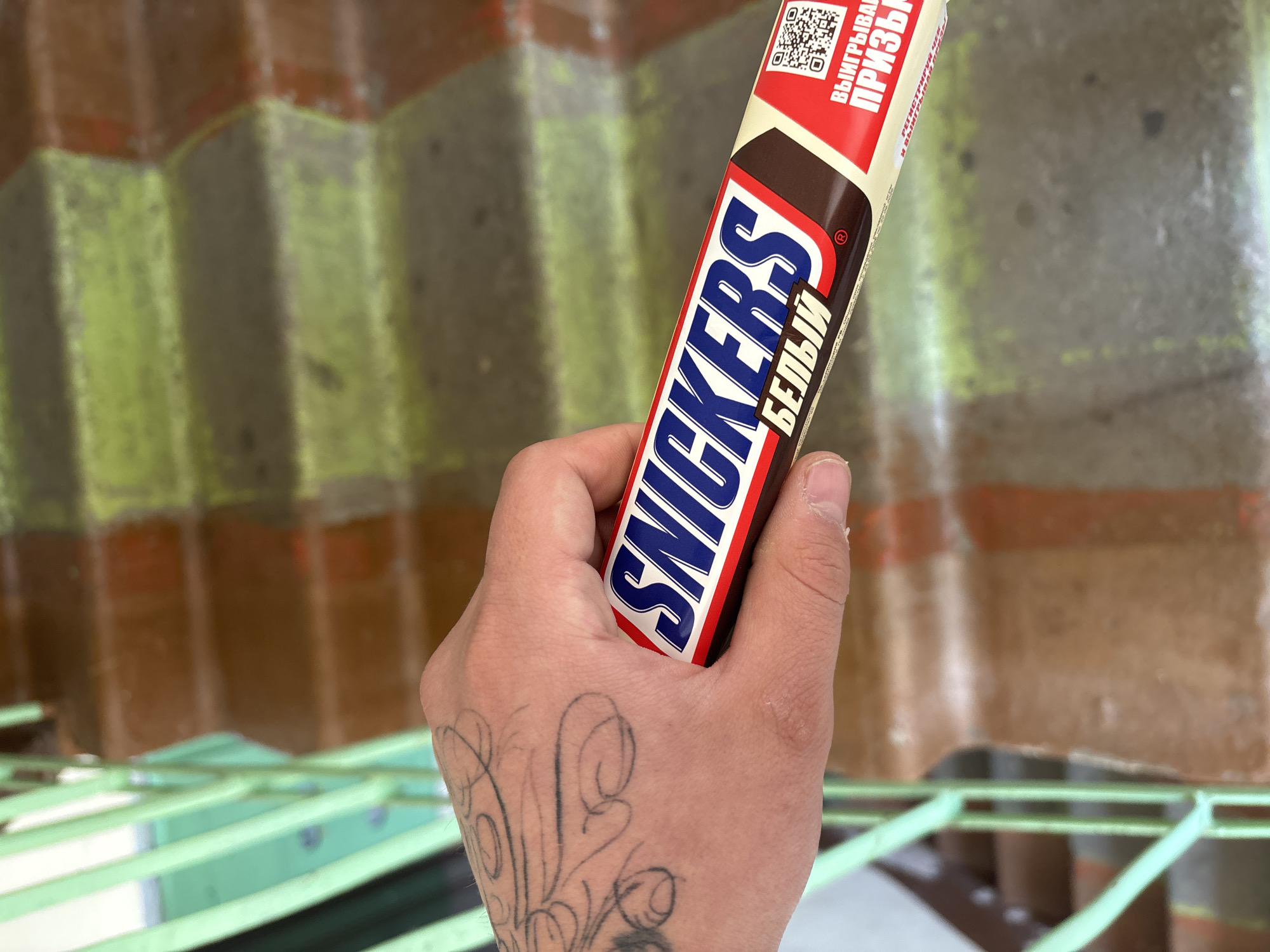 A hand with a tattoo holds a "Snickers Белый" candy bar in front of a brown and green corrugated wall. The location is unknown.