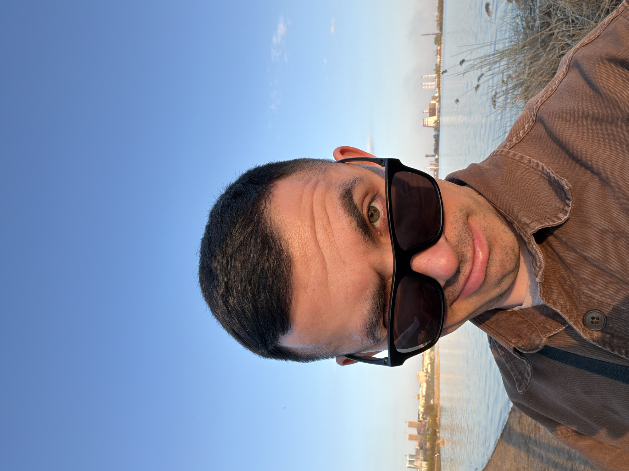 A man with dark hair and sunglasses poses for a selfie outdoors. He's wearing a brown shirt with buttons. Behind him is a body of water with a city skyline and industrial structures visible in the distance. Reeds grow along the shoreline. The sky is blue. Given the industrial buildings in the distance and the reeds on the bank of the water, this image was likely taken in Izhevsk, Russia near the Izhevsk reservoir.
