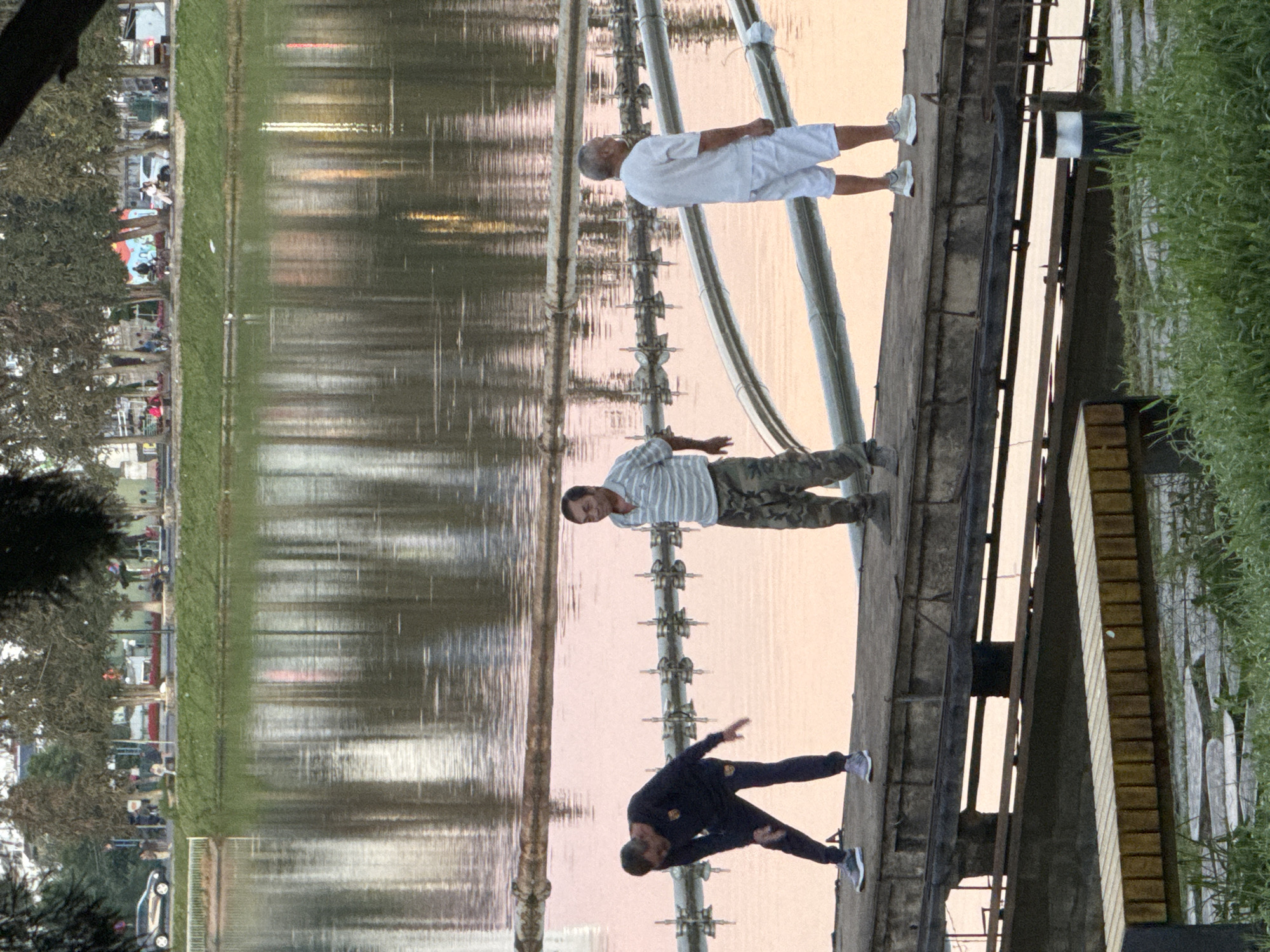 The image depicts an outdoor scene at a body of water, likely a lake or reservoir. Three men are standing on a concrete pier. The man on the left is wearing a dark long-sleeved shirt and dark pants, and appears to be stretching. The man in the middle is wearing a striped short-sleeved shirt and camouflage pants, and is looking towards the left. The man on the right is wearing a white short-sleeved shirt and white shorts, and is looking to the right. In the background, there is a reflection of trees and buildings on the water. The overall atmosphere suggests a relaxed, possibly early morning or late afternoon setting. The presence of industrial-looking equipment, possibly related to water treatment or aeration, is visible along the pier's edge. The scene is not specifically identifiable as being in Da Lat, Vietnam without additional contextual information.