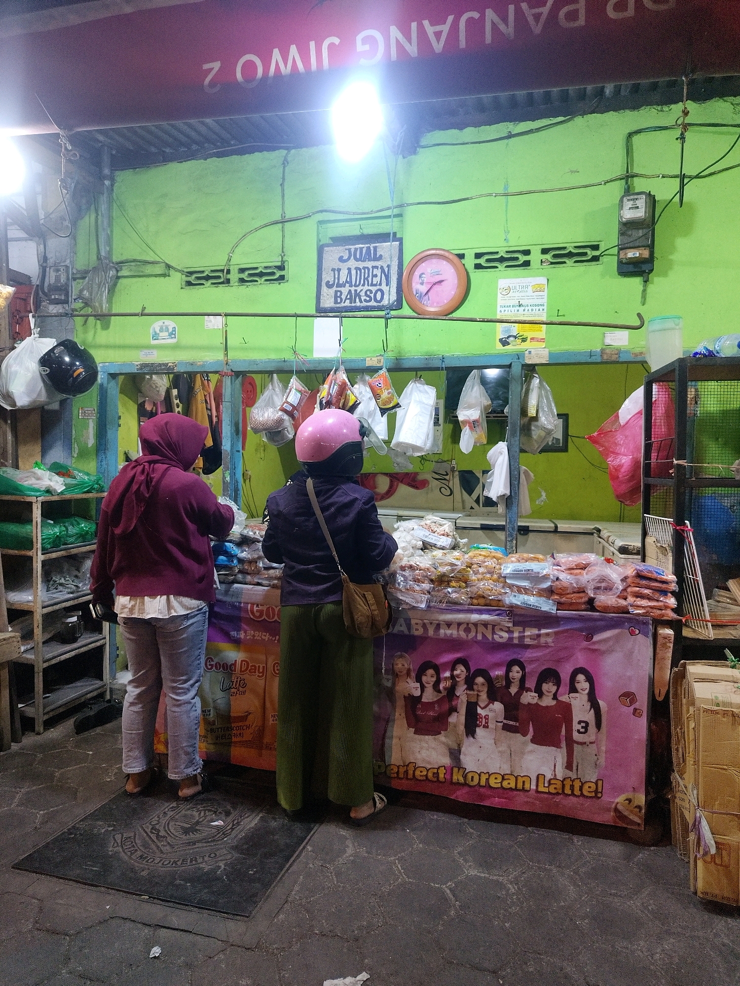 The image depicts a street-side food stall in Mojokerto, Indonesia, specializing in "Jajanan Bakso" (fried snacks and meatballs). The stall is adorned with a vibrant green wall and a maroon banner that reads "JUAL Jladren Bakso". Above the stall, a red awning with white lettering is partially visible, showing "PANJANG JIW0 2". Two women are seen at the stall, one in a maroon hijab and a white collared shirt, the other in a pink helmet and dark clothing. They appear to be customers or staff members, engaged in selecting or preparing food items.
The stall is laden with various packaged food products, including what appear to be sausages, fried snacks, and other processed foods. Some items are displayed in plastic bags and on shelves. A large banner at the front of the stall features a group of young women and the text "BABYMONSTER" and "Perfect Korean Latte!". Behind the stall, there are refrigerators and shelves stocked with more merchandise. To the left, a black motorcycle helmet is hung, and to the right, a wire cage structure is visible. The ground is paved with interlocking stones, and a dark, circular plaque with an emblem and text that reads "KOTA MOJOKERTO" is embedded in the pavement in front of the stall. The overall scene suggests a lively local market or street food area, bustling with activity.