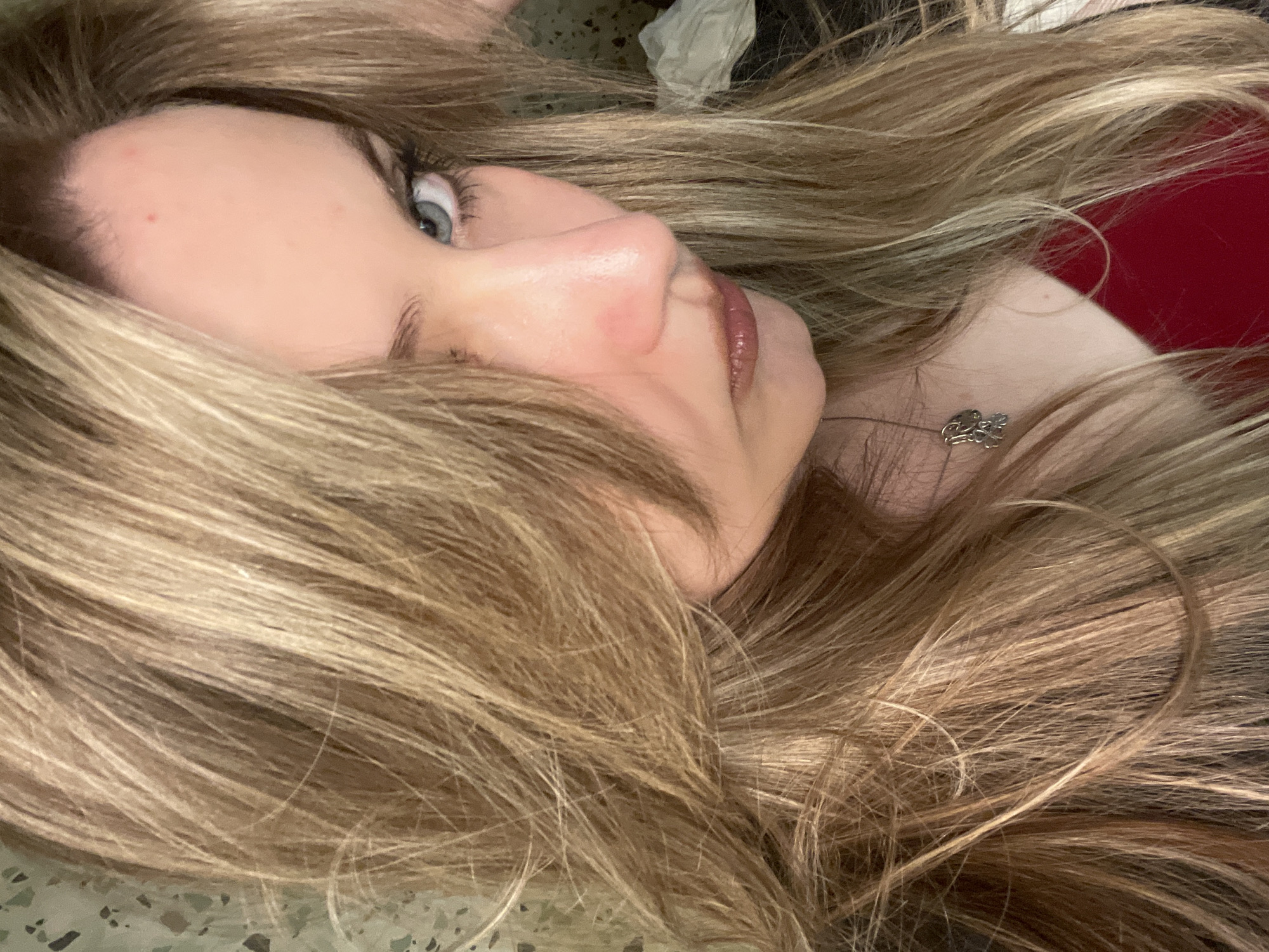 A close-up, low-angle, selfie-style shot focuses on the face and upper torso of a woman. She has fair skin, blue eyes, and long, blonde hair that is slightly windblown and partially obscures her face. Her expression is neutral, with her lips slightly pursed. She is wearing a red top and a silver necklace with a small, abstract pendant. The background appears to be an indoor setting with a patterned wall on the left and a crumpled white object on the right. The lighting is soft, with some glare on her skin.