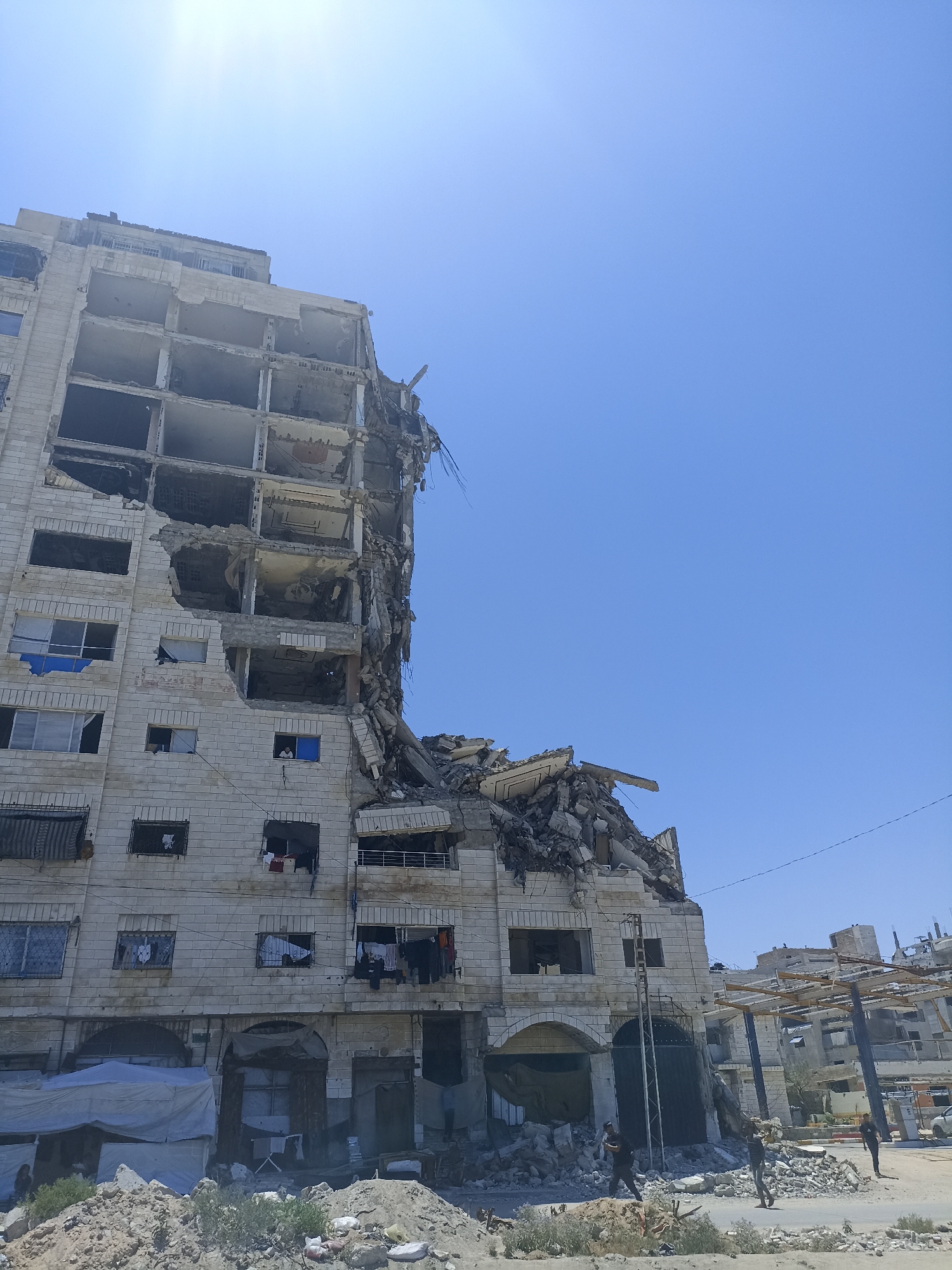 A partially destroyed apartment building stands under a bright, clear blue sky. The building, constructed of light-colored bricks, has suffered significant damage, with sections of its facade and interior structure exposed. Many windows are missing or broken, and debris is piled at the base of the building and scattered in the foreground. The top floors of the building are heavily damaged and appear to be collapsing.
In front of the building, there are several makeshift structures, possibly tents, made of tarpaulin. Some of these structures have clothing hanging from them. The ground in front of the building is covered in rubble, sand, and some sparse vegetation.
In the middle ground, a few people are visible near the damaged building and the rubble. One person is walking towards the camera, and two others are further away, one of them appearing to be carrying something. The scene suggests the aftermath of a destructive event, possibly an attack or a natural disaster, given the extensive damage to the building and the presence of rubble.
In the background, more damaged buildings and structures are visible, suggesting a wider area of destruction. There are some overhead wires or cables in the scene, and what appears to be a road or pathway in the foreground and middle ground. The sun is high in the sky, indicating it is likely daytime, possibly midday, and the weather appears to be clear and sunny. There is no visible text in the image.