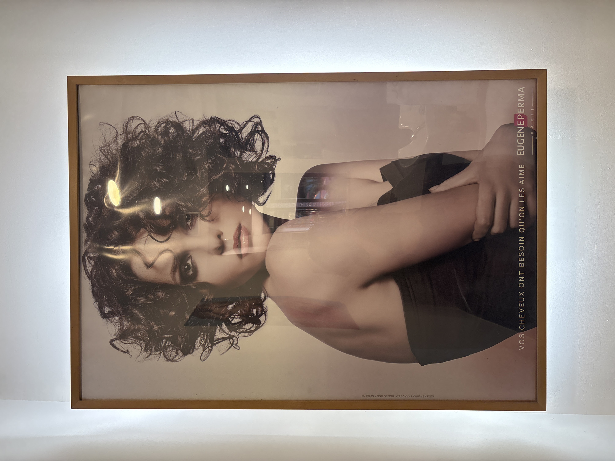 An advertisement poster featuring a woman with curly brown hair is hanging on a white wall. The poster is framed in wood and is lit from the front, causing reflections on the glass surface. The woman is looking directly at the viewer, with her arms crossed. The poster contains text in French, including the slogan "VOS CHEVEUX ONT BESOIN QU'ON LES AIME" (Your hair needs to be loved) and the brand name "EUGENE PERMA PARIS". The background is a plain, light color. The poster appears to be located in a public space, possibly a salon or a shop, given the nature of the advertisement. Without further visual clues, it is impossible to determine the specific location within the city of Rome, Italy.