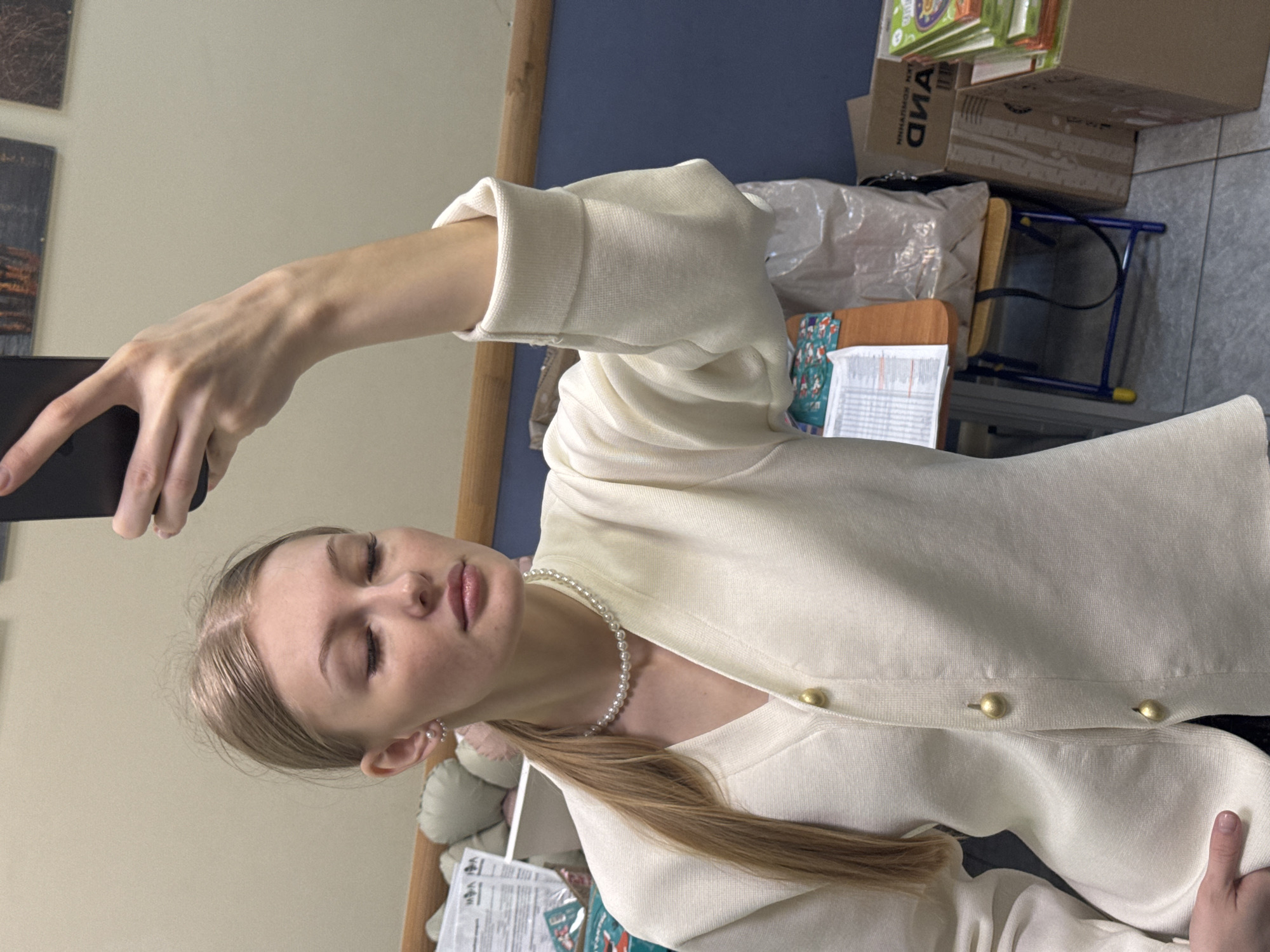 A young, fair-skinned woman with long blonde hair is taking a selfie indoors. She is wearing a white cardigan with gold buttons and a pearl necklace. The background is slightly cluttered with furniture and boxes. The location cannot be identified as being in Perm, Russia based on this image alone.