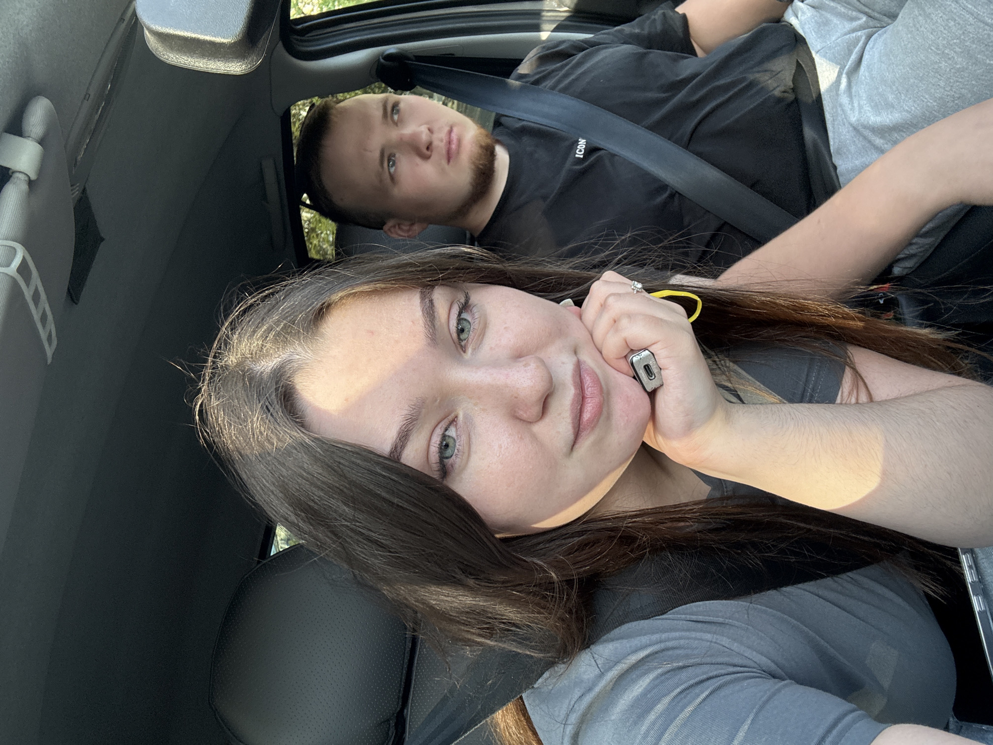 The image shows two people, a woman and a man, in the front seats of a car. The woman is in the foreground, facing the camera, with her left hand supporting her chin. She has long brown hair and is wearing a grey t-shirt and a seatbelt. The man is in the background, looking to the right. He has short brown hair and a beard, and is wearing a black t-shirt and a seatbelt. The interior of the car is visible, including the dashboard and the seats. The lighting suggests it is daytime. The location is identified as Perm, Russia.