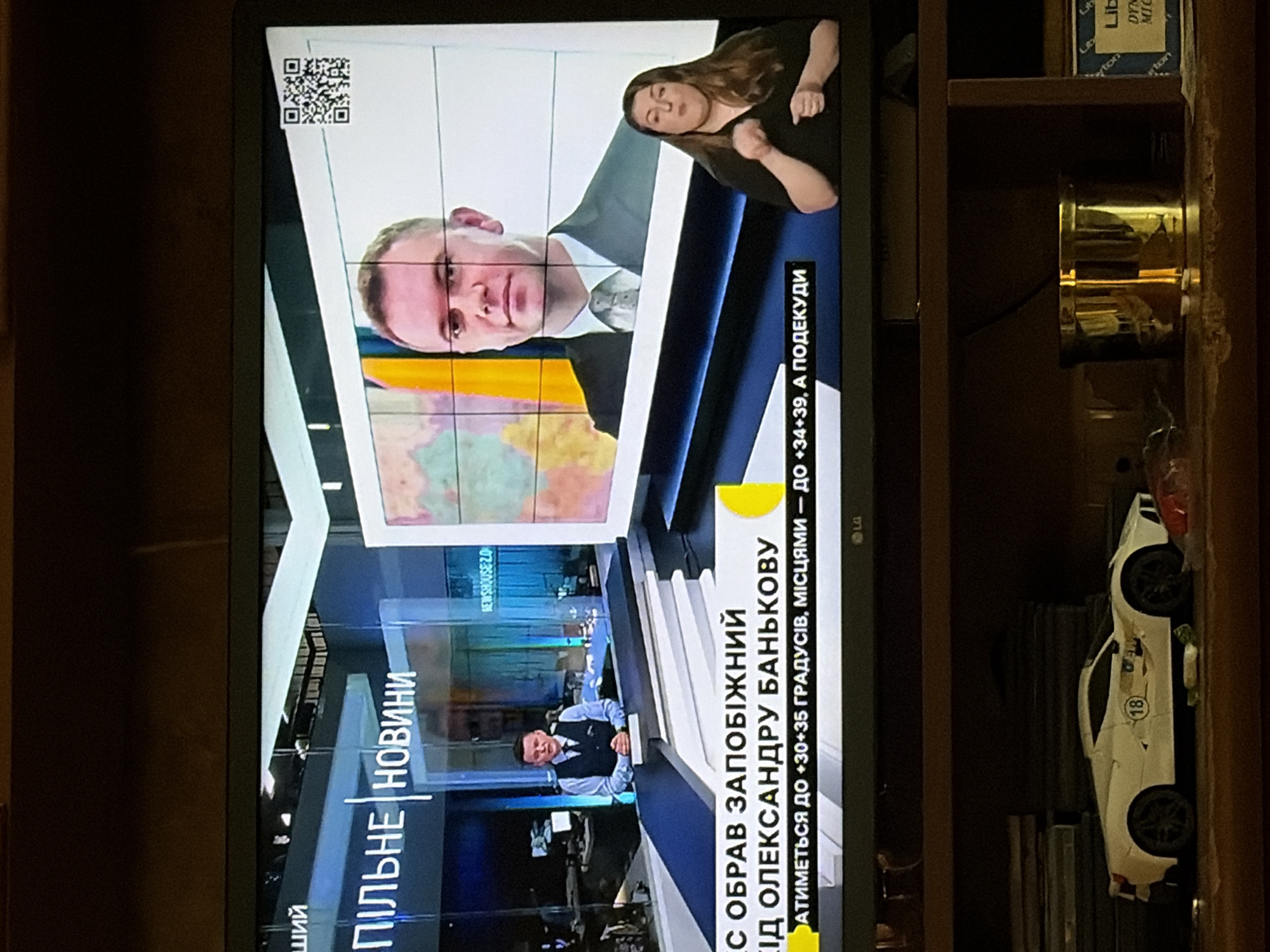 The image shows a television screen displaying a news broadcast. In the foreground, a white toy sports car is positioned on a shelf. The news broadcast features a male anchor at a desk on the left side of the screen and a sign language interpreter on the right. Behind them, a large screen displays a map-like image and a QR code. Text overlays on the screen indicate "ПІЛЬНЕ НОВИНИ" (Urgent News) and details about the weather. The background of the room is dark.