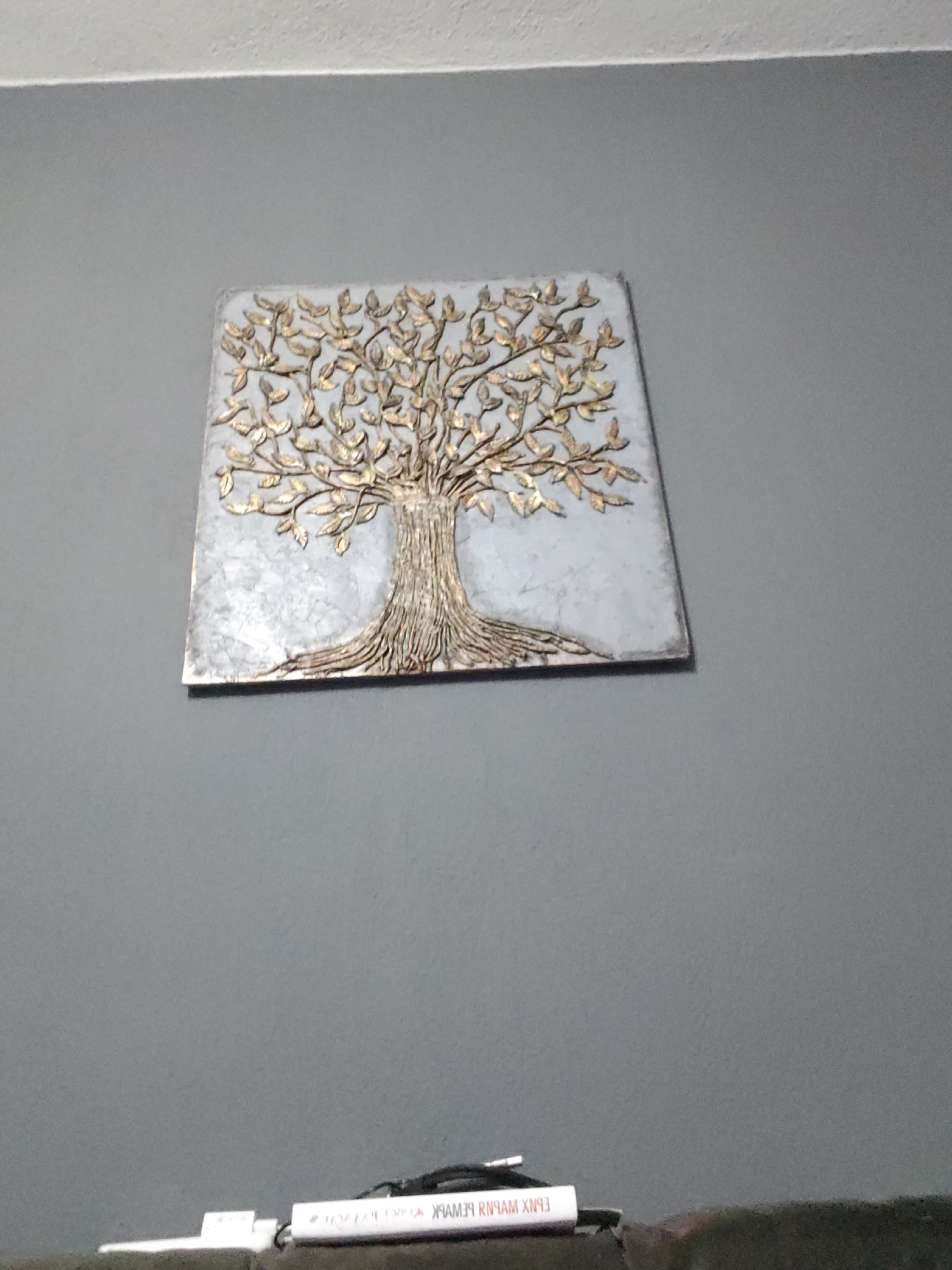 A tree of life artwork hangs on a gray wall. Below it is a white object with black wires.