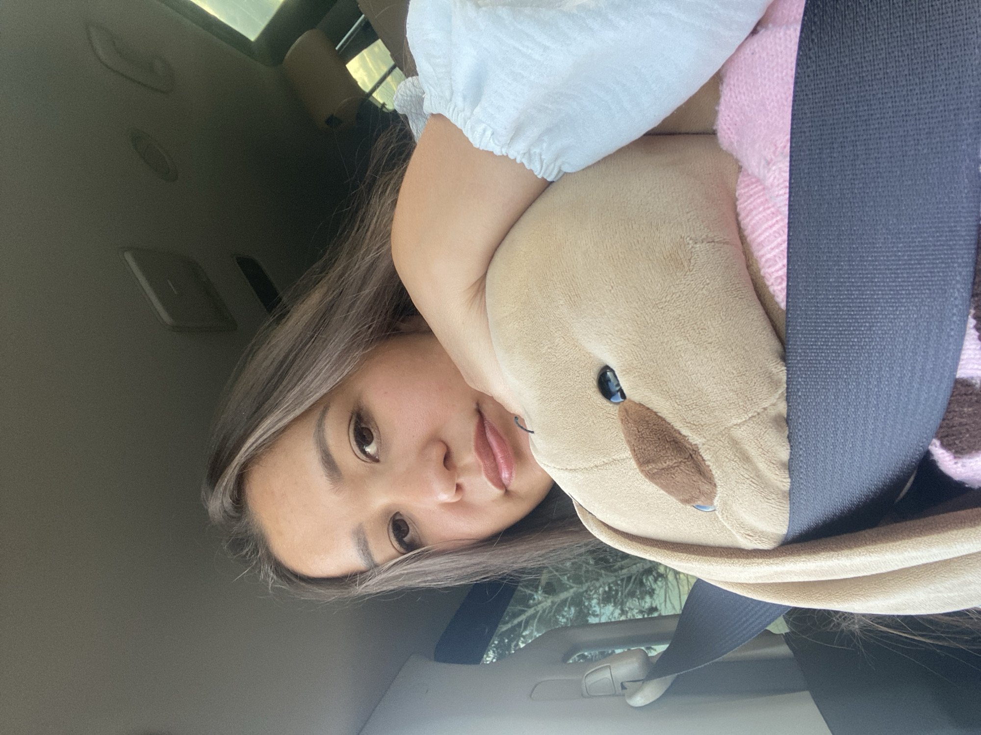 A woman is in the passenger seat of a vehicle, holding a stuffed animal. She is wearing a light-colored top with ruffled sleeves. The stuffed animal is brown and appears to be a bunny. A seatbelt is visible across both the woman and the stuffed animal. The background shows the interior of the car, including the ceiling and a portion of the driver's seat. Outside the car window, trees are visible. The lighting suggests it is daytime. The location of Staromusino, Russia is not visible or inferable from the image.