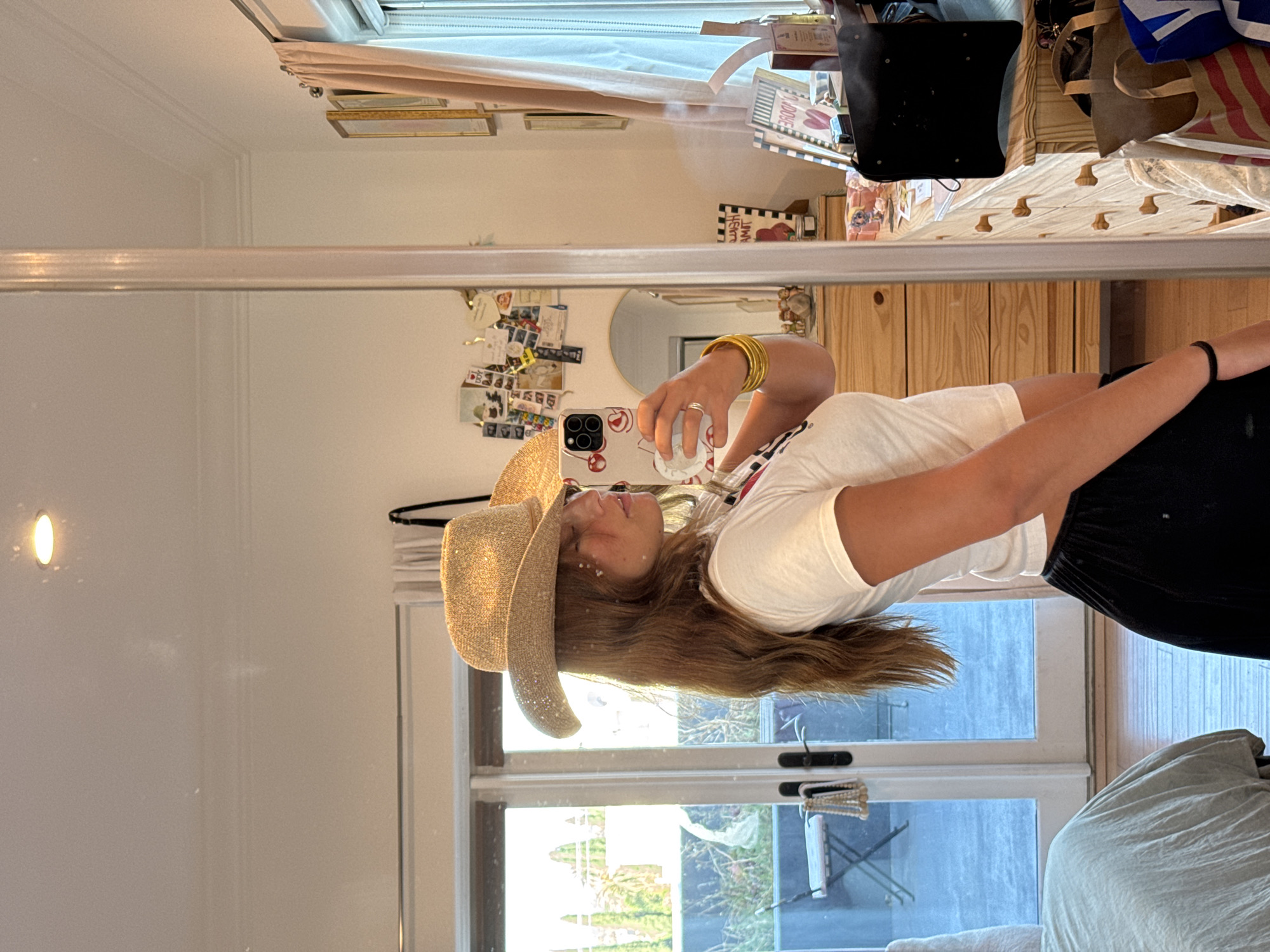 A woman wearing a straw cowboy hat and a white t-shirt is holding a smartphone with a cherry-patterned case in front of a mirror. She is wearing multiple gold bracelets on her wrist. The mirror reflects a room with various items on shelves, including books and bags. Outside the window, trees and possibly buildings are visible. The location is not specified in Los Angeles.