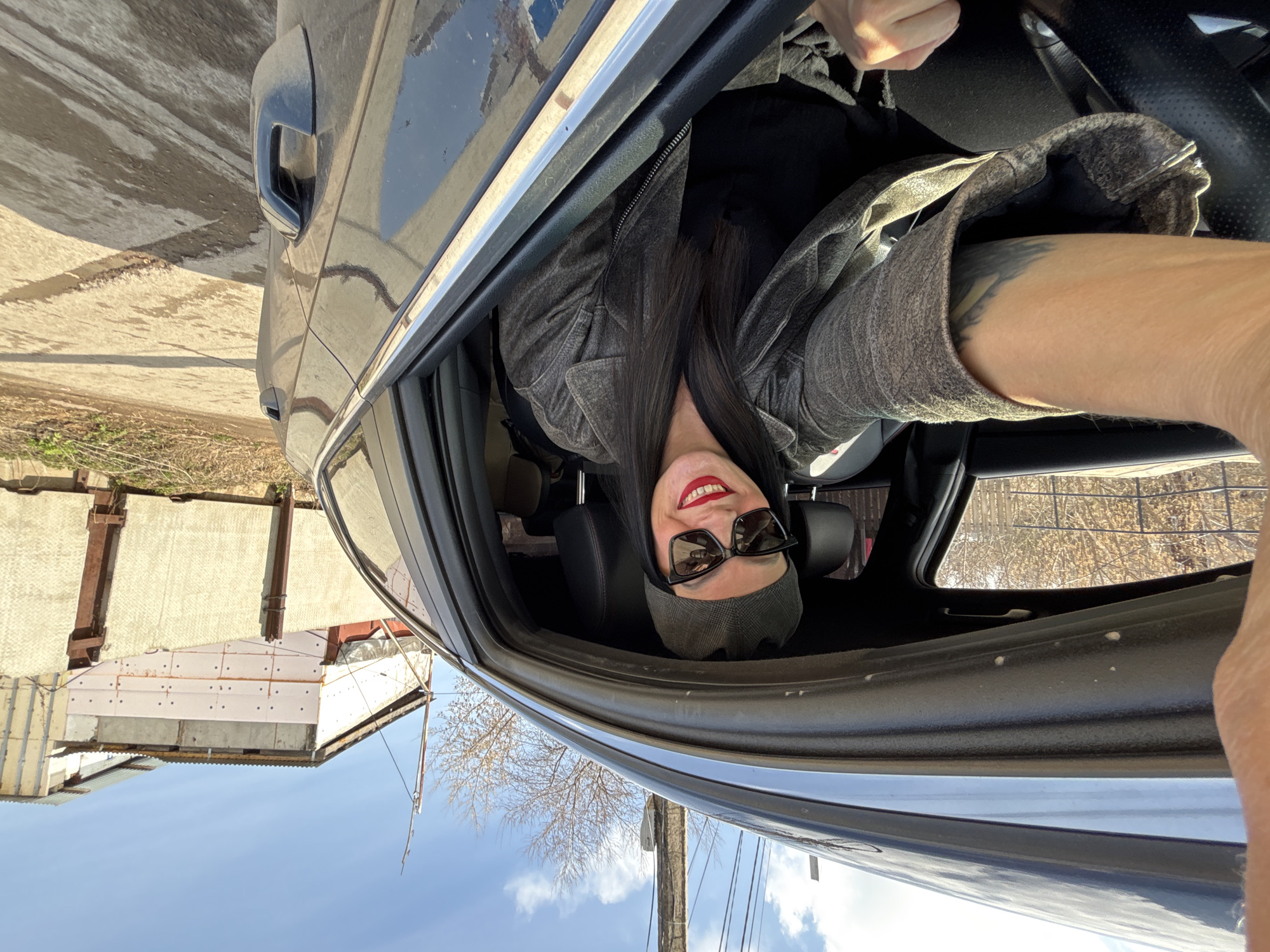 A woman is inside a car, possibly a selfie from the driver's seat, viewed from an unusual angle. She is wearing dark sunglasses, bright red lipstick, and a gray hat. She also has a gray jacket with a black shirt underneath. The woman's dark hair falls past her shoulders. Her left arm, visible with a tattoo, is extended towards the camera. The car appears to be parked on a street in the city of Chaikovsky, Russia. The background includes a building with a rusted roof, a utility pole, power lines, and a blue sky with some clouds.