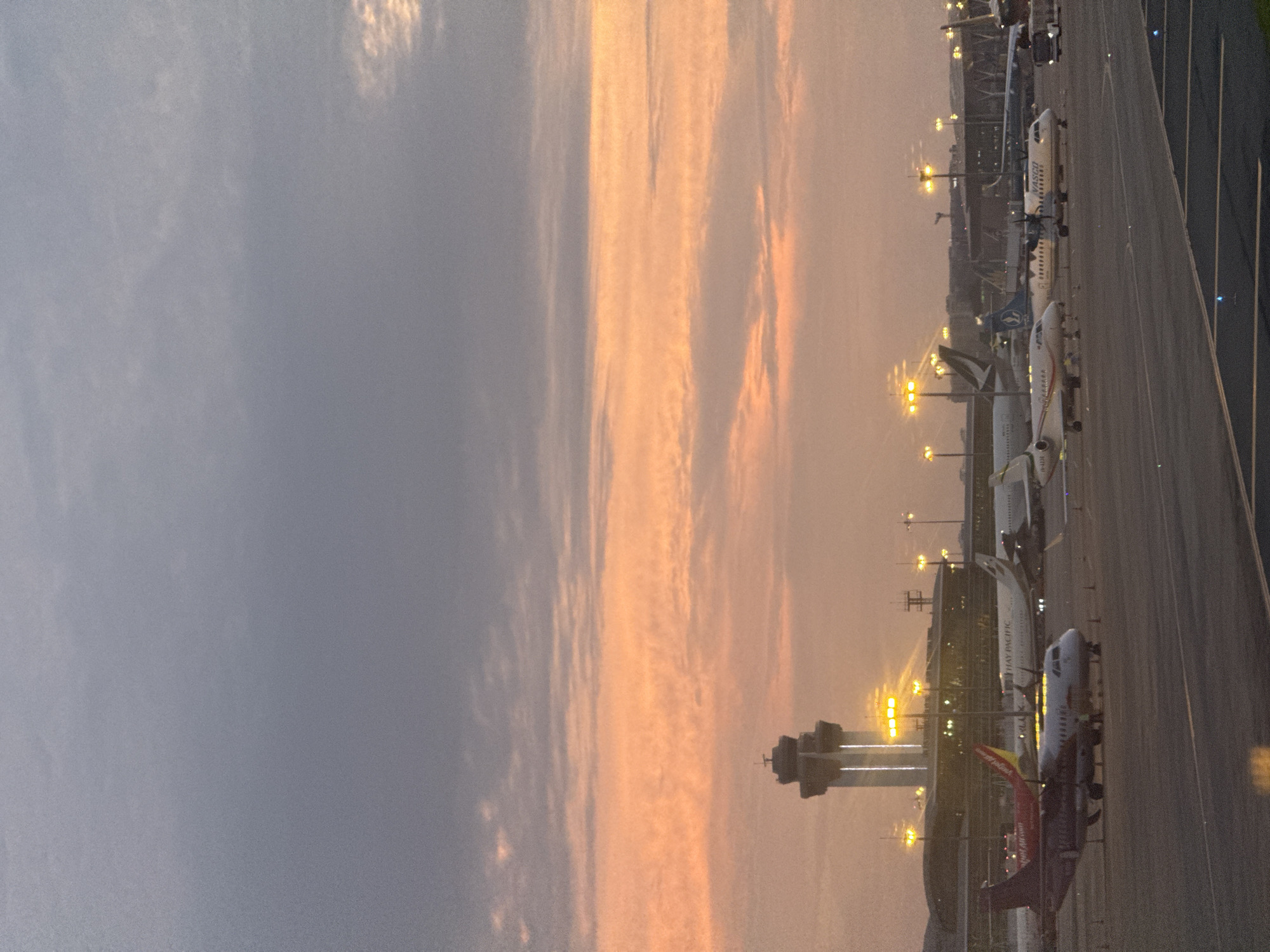 At dusk at Tan Son Nhat International Airport in Ho Chi Minh City, Vietnam, several commercial airplanes are parked on the tarmac. In the foreground, a red and white passenger jet with the logo "Vietjet Air" is visible. Further back, a larger aircraft with the "Cathay Pacific" logo is parked. An air traffic control tower, illuminated by bright lights, stands tall in the background against a sky painted with hues of orange and gray from the sunset. The airport is bustling with activity, as indicated by the presence of ground crew and service vehicles near the aircraft.