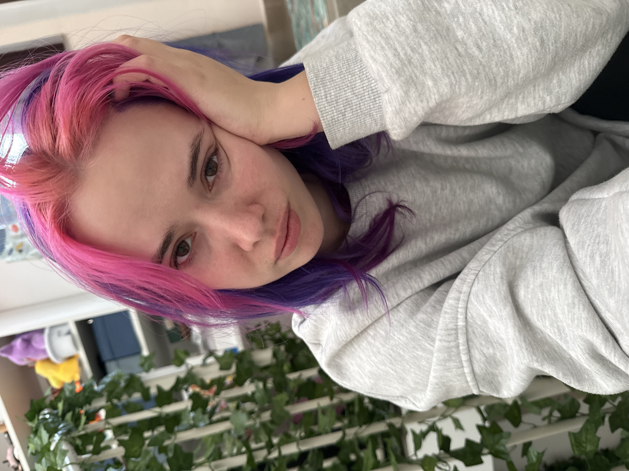 A young woman with vibrant pink and purple hair is the focal point of the image. She wears a light gray hooded sweatshirt and rests her head on her hand. The background features green ivy-like plants, adding a touch of nature to the indoor setting. The image appears to be taken in a room with hints of other objects in the blurred background, such as furniture and possibly a shelf. There is no information about the location in the city of City of Zagreb, Croatia.