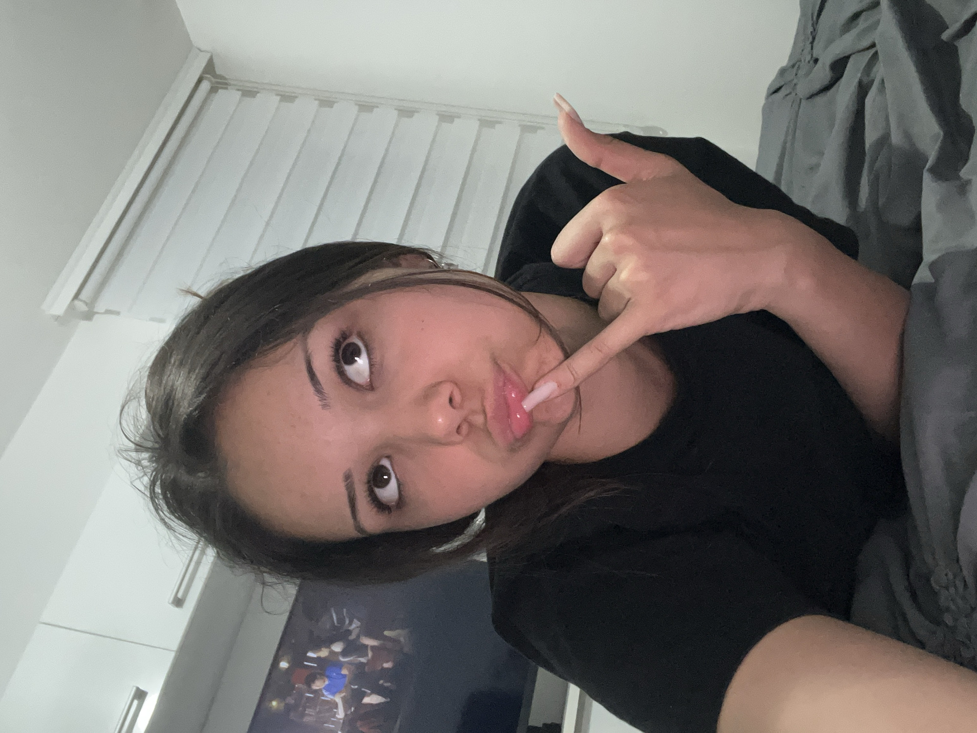 A young woman with dark hair, wearing a black t-shirt, lies on a grey bed. She is looking upwards with her finger to her lips, and her right hand is positioned as if making a "shaka" sign. Behind her, a television screen displays an image of people. To her right, a white horizontal blind is visible on the wall. The environment appears to be an indoor bedroom setting.