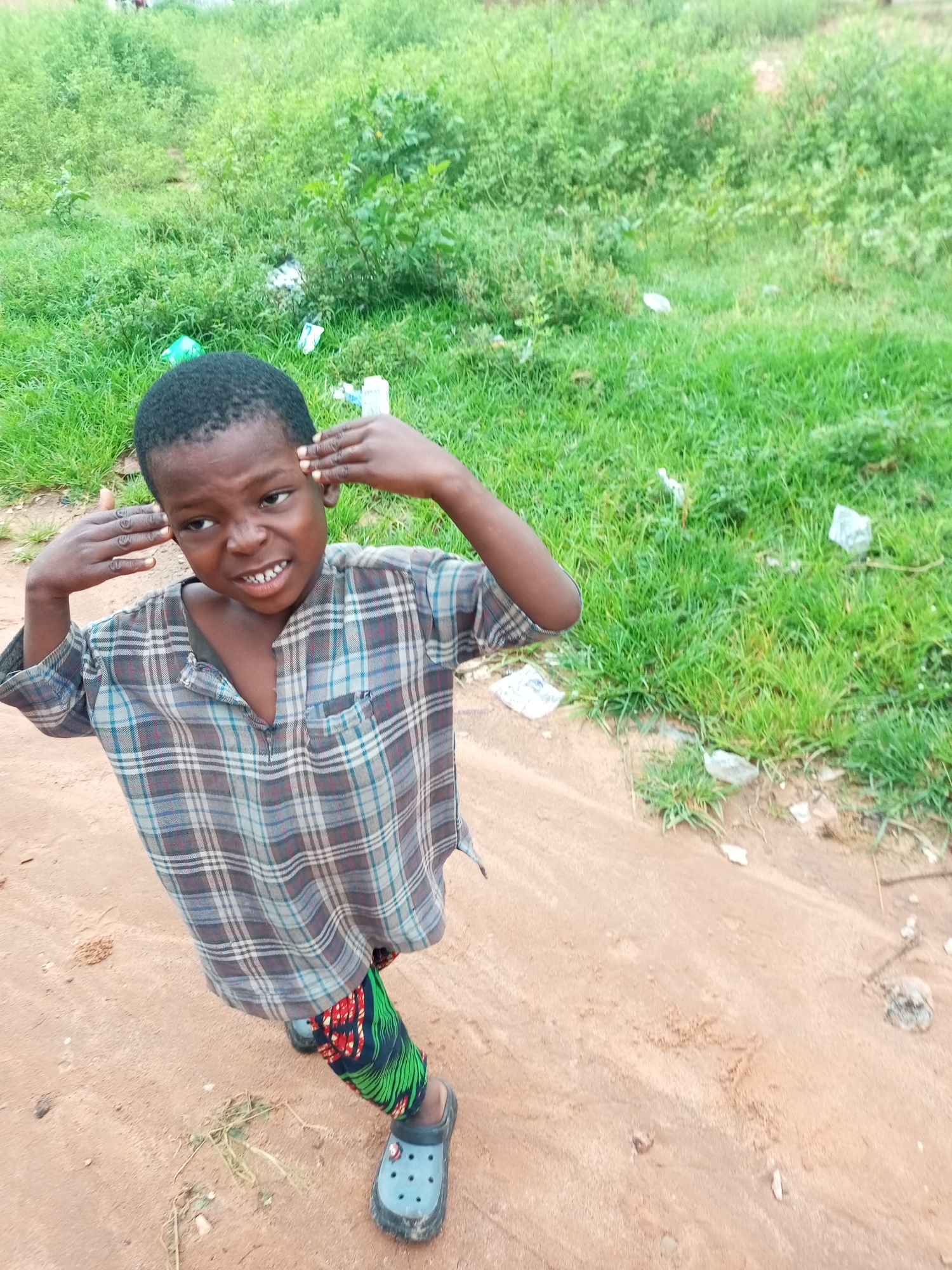 The image, taken in Tempure, Nigeria, shows a young boy standing on a dirt path with lush green foliage and some scattered litter in the background. The boy, who appears to be of African descent, is wearing a plaid shirt and vibrantly patterned green and red pants, along with blue Croc-style shoes. His hands are positioned on the sides of his head, and he has a somewhat pensive expression, with his mouth slightly open revealing his teeth. The lighting suggests it is daytime, and the environment appears to be a semi-rural or undeveloped area.