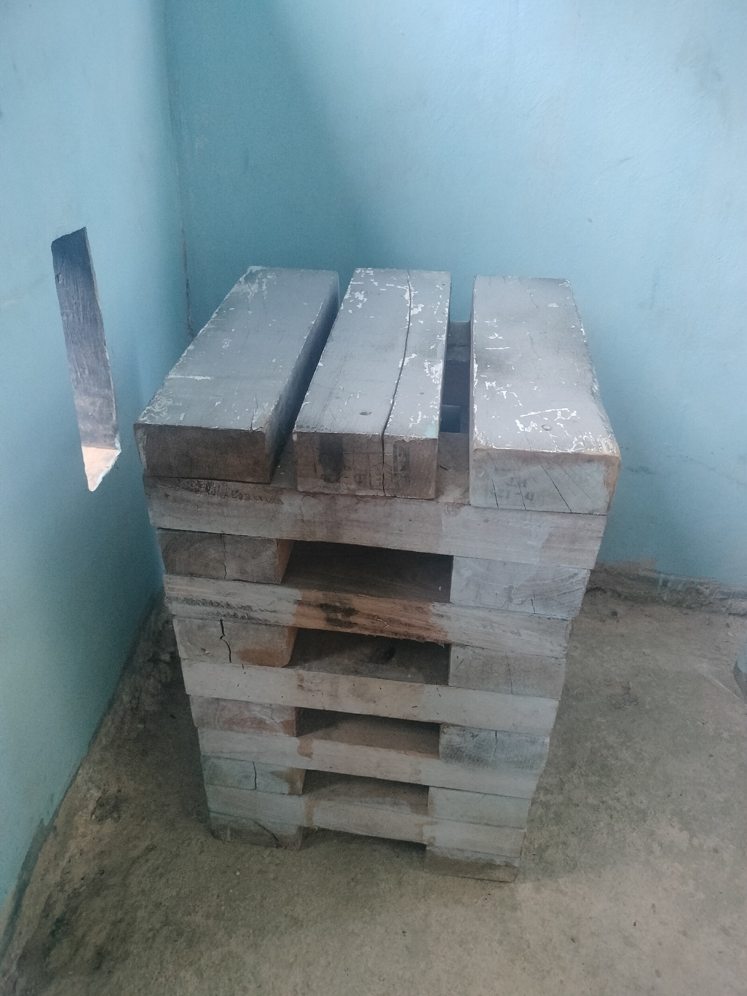 An outdoor scene in Batam, Indonesia, features a stack of wooden pallets leaning against a light blue wall. The pallets are weathered and appear to be grey. A narrow, irregularly shaped opening is visible in the wall to the left of the pallets. The ground beneath the pallets is rough concrete or dirt. The lighting suggests it may be daytime.