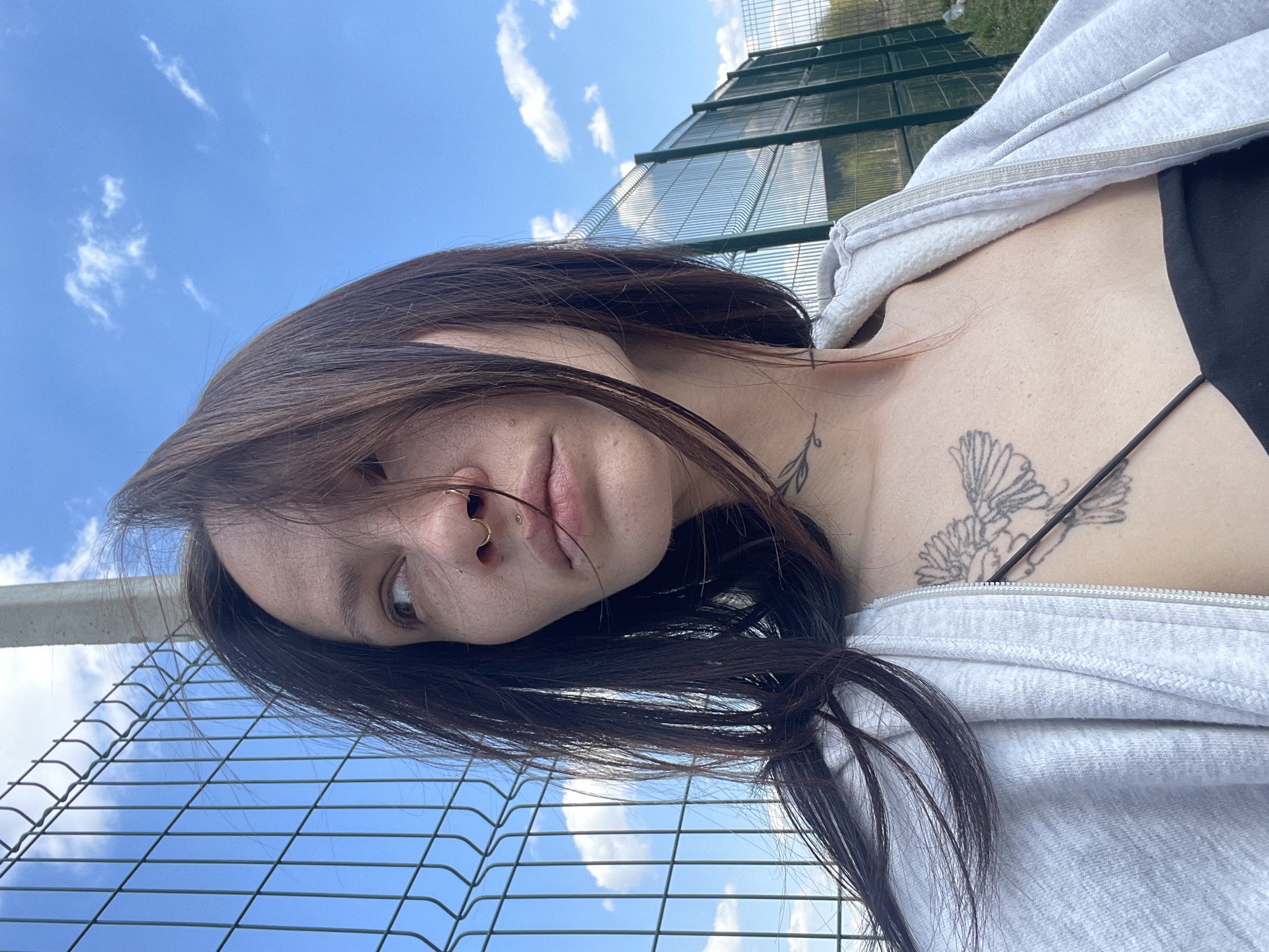 A medium shot of a young woman outside, possibly in Izhevsk, Russia, given the location request. The woman has long brown hair and is wearing a light gray zip-up hoodie over a black top. She has a nose piercing and tattoos on her chest and neck. She is positioned close to a chain-link fence, with a similar fence structure visible in the background along with greenery. The sky is blue with scattered clouds. The image is rotated, creating a tilted perspective.