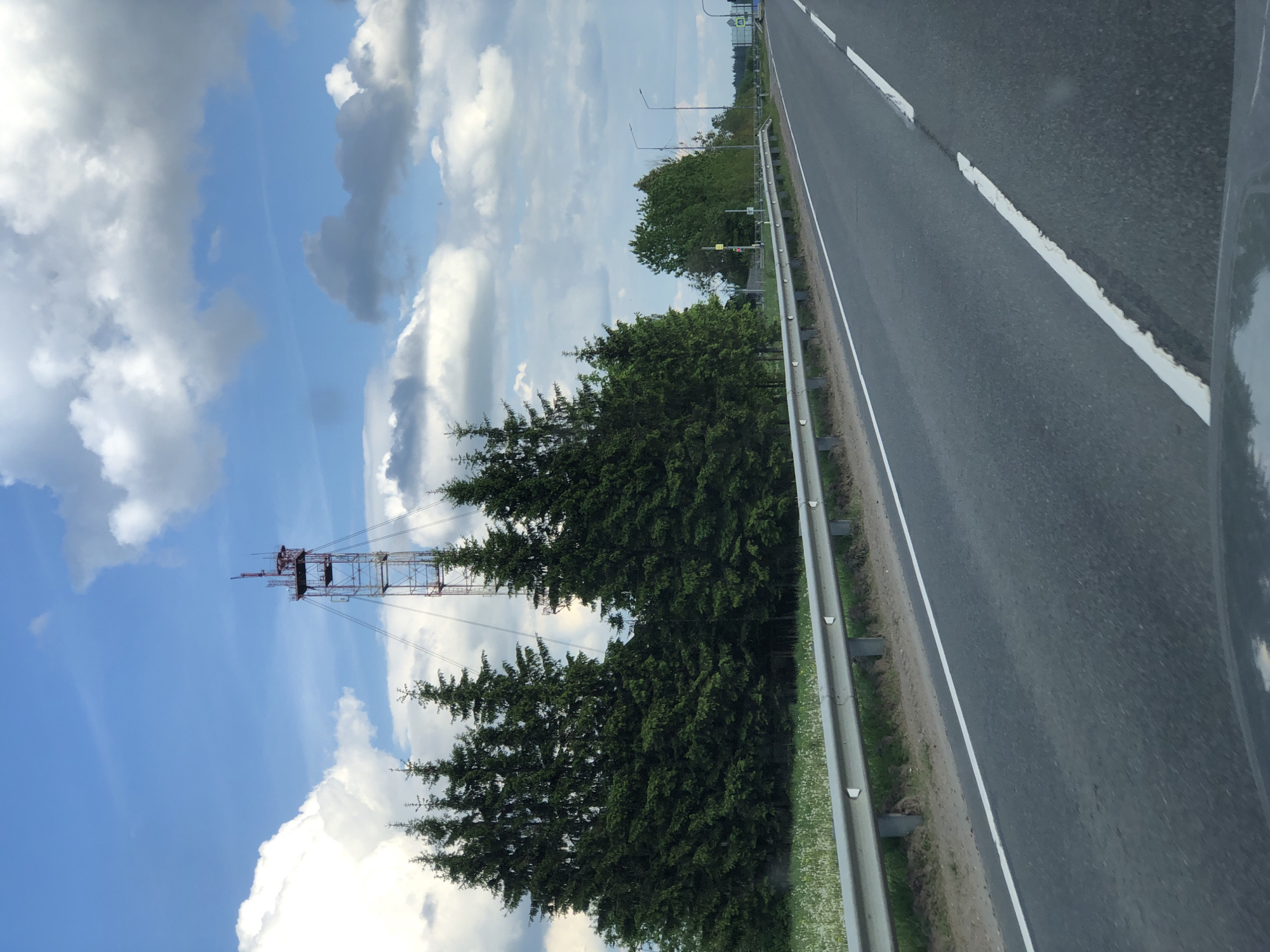 This media file captures a daytime scene along a road in Dyatlitsy, Russia, likely from a moving vehicle. The image is oriented vertically, with the road stretching from the lower right towards the top center.
The scene features a multi-lane asphalt road with clear white markings, including a solid line on the shoulder and a broken line separating traffic lanes. A metal guardrail runs along the left side of the road (as pictured), separating it from a strip of green grass interspersed with small white flowers. Beyond the guardrail, and extending across the left side of the frame, are dense rows of dark green coniferous trees, possibly spruces or pines.
Dominating the upper left portion of the image, rising above the treeline, is a tall, red and white metal lattice tower. This communication or radio tower is equipped with various antennas and structural elements at its apex, with wires extending from it.
The sky occupies the majority of the background, displaying a bright blue hue with scattered white and grey cumulus clouds. The lighting suggests it is mid-day or afternoon, and the weather appears fair. No people or legible text are visible in the frame. The overall impression is one of a highway or main road passing through a forested, possibly rural, landscape.