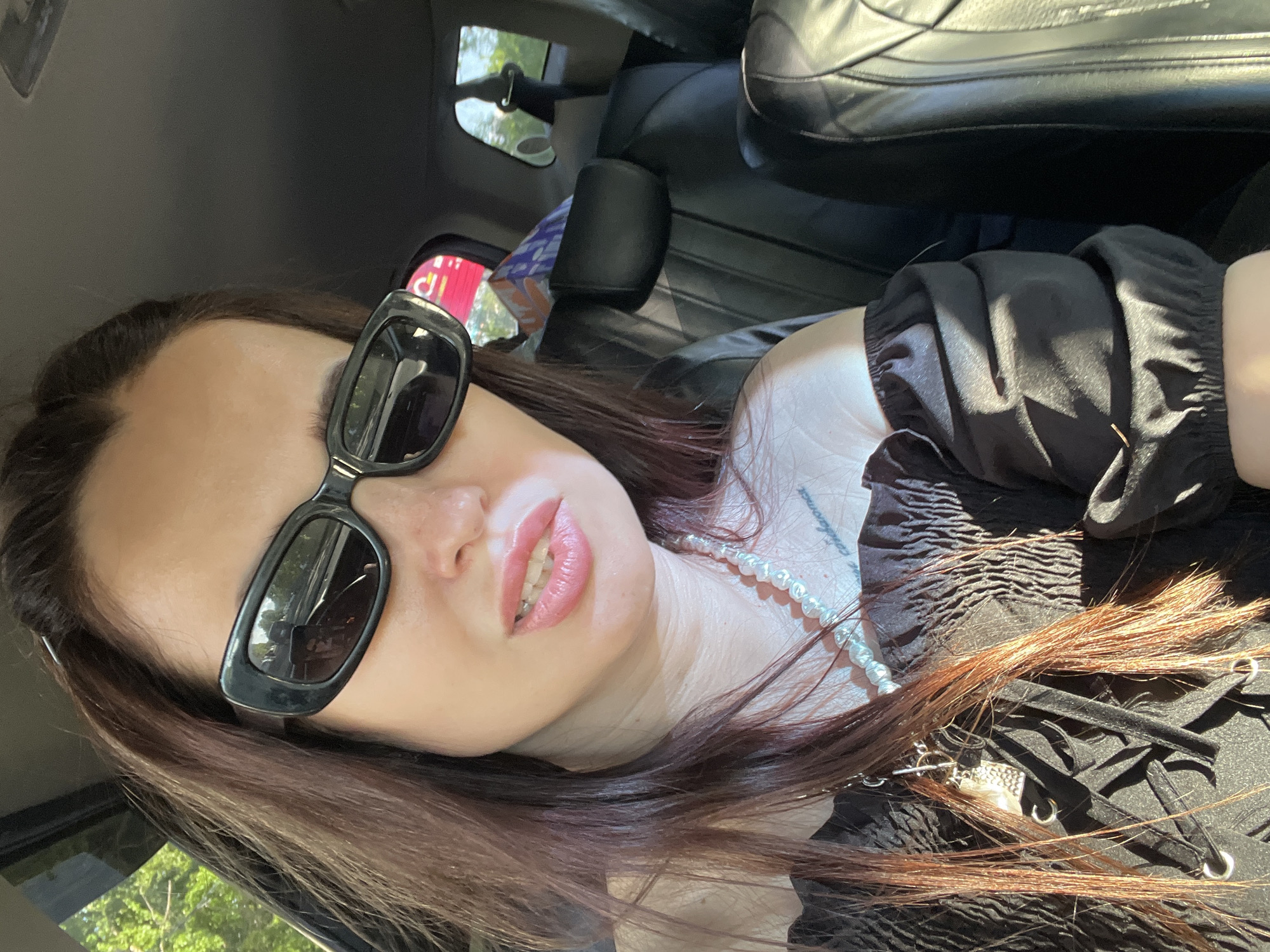 A young adult female is depicted in a close-up selfie, taken inside a car. The image is oriented sideways. She has long, dark brown hair with reddish highlights. She is wearing large, black, rectangular sunglasses that cover her eyes, and her lips are painted with pink lipstick or gloss, slightly parted to reveal her teeth. Around her neck, she wears a light-colored, possibly pearl, necklace.
Her attire consists of a black, off-the-shoulder top featuring ruffled sleeves and a smocked bodice. The front of the top has a corset-style lacing with small silver eyelets. A tattoo with faint text, possibly starting with "const", is visible on her upper right shoulder/collarbone area.
The background shows parts of the car's interior, including dark leather seats and a dark headliner. Bright sunlight streams in, creating highlights on her hair and clothing. Through the car windows and reflected in her sunglasses, green foliage from trees and a bright sky are visible, indicating it is daytime. A faint red object with indistinct white markings, possibly a sign, is reflected in one of the sunglass lenses. The subject's expression is neutral, suggesting a posed moment.
