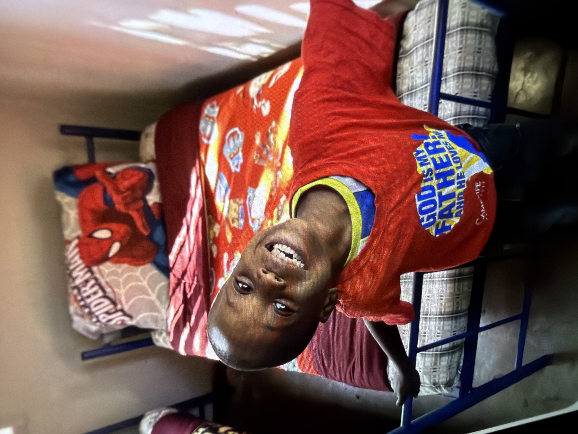 A young boy, with dark skin and a wide smile, is leaning towards the camera. He is wearing a red t-shirt with yellow and blue lettering that reads "GOD IS MY FATHER AND HE LOVES ME." The boy is positioned in front of a twin-sized bed with blue metal bed frame. The bed is covered with a red blanket featuring cartoon characters and a Spiderman pillow. The scene appears to be indoors, with a plain wall in the background and light casting shadows. The boy's left hand is resting on the bed frame. The setting is consistent with a bedroom, likely in a child's room.