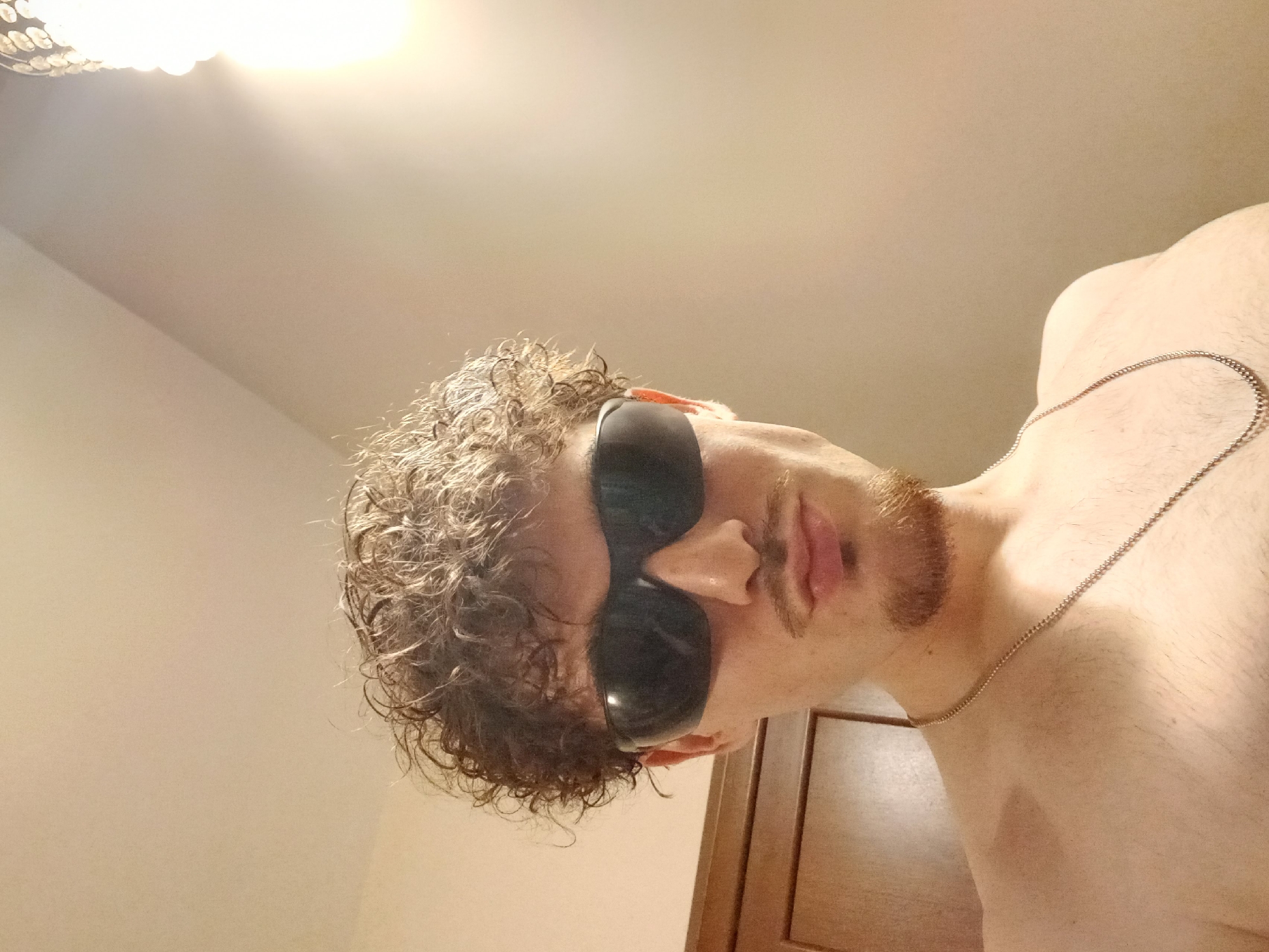The image shows a young man with curly brown hair and a goatee wearing sunglasses and a silver chain necklace. He is shirtless and looking directly at the camera. The background is a plain wall with a light fixture on the right side. A wooden door is visible on the left side of the image. There is no information available to suggest this image is located in Parvomay, Bulgaria.
