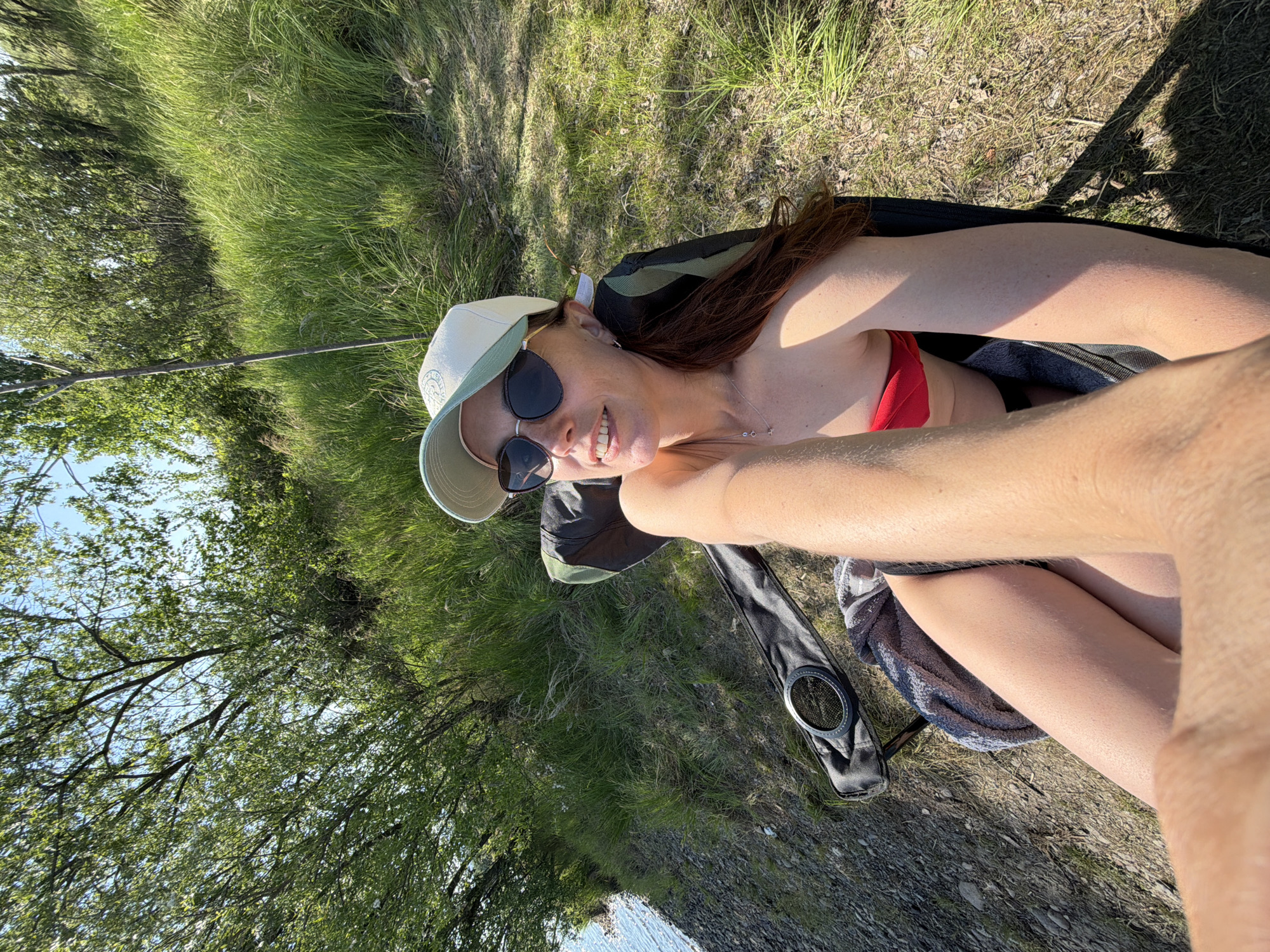 A woman wearing sunglasses, a white cap, and a red bikini top sits on the ground near a grassy area with a towel and a black case next to her. There are trees in the background and the edge of water can be seen. The location is near a body of water, possibly in a park or recreational area in Birobidzhan, Russia. The scene suggests a relaxing outdoor activity.