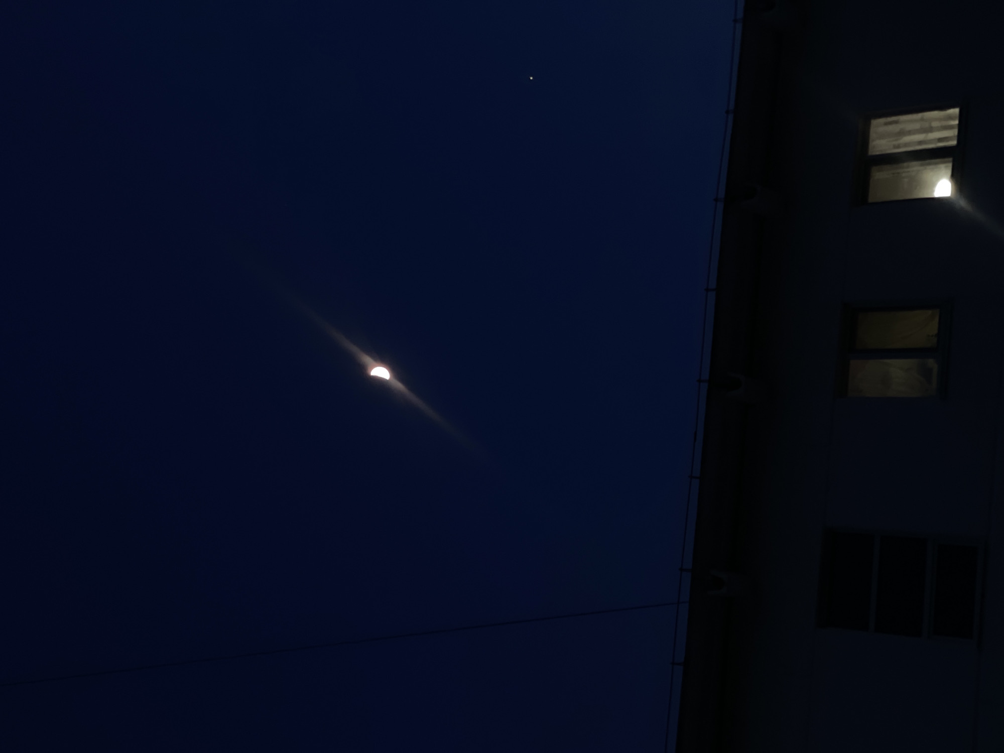 An outdoor, low-light photograph captures a night sky over Chaikovsky, Russia, alongside a section of an urban building. The sky is a deep, dark blue, indicating either late evening or early morning twilight, and appears clear. A bright crescent moon, likely in its first or last quarter phase, is prominent in the lower left portion of the sky, surrounded by a soft glow or light trail. Above and slightly to the left of the moon, a solitary, very bright star or planet is clearly visible as a distinct point of light.
A light-colored, possibly off-white or light grey, building occupies the right side of the frame. Several rectangular windows are visible on its facade. On an upper floor, one window on the far right glows brightly from an internal light source, creating a visible reflection. Below it, another window appears darker but might also show a faint interior light. Further down on the building, three smaller, dark windows are visible. A ladder-like structure or a series of pipes runs vertically along the left edge of the building. A thin, dark line, possibly a power line or wire, traverses the lower mid-ground of the sky horizontally, passing behind the moon. No people or text are visible, and the scene conveys a quiet, tranquil urban night atmosphere.