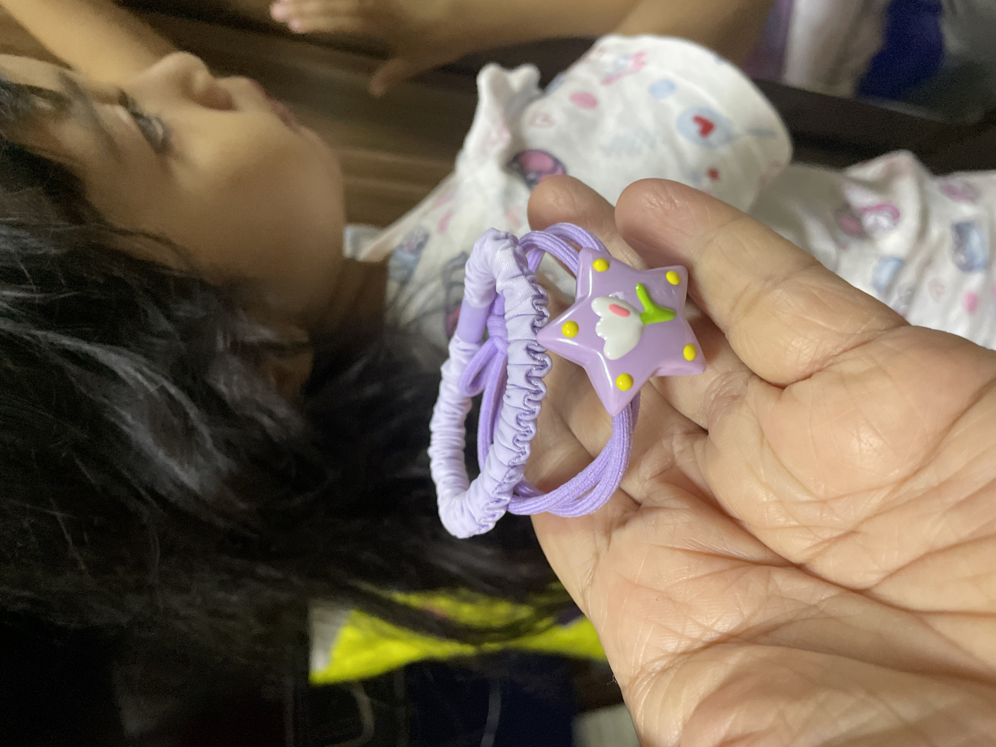 An indoor, eye-level shot in Davao City, Philippines, captures a moment between a child and an adult. In the foreground, an adult's hand, characterized by visible skin creases, holds two purple hair accessories. One is a plain elastic hair tie, and the other is a thicker, ruched elastic band. Attached to the plain elastic is a light purple, star-shaped charm decorated with yellow dots and a white and green tulip-like design.
The child, with dark, wavy hair, is partially visible in the mid-ground, lying down on what appears to be a bed or couch. She is wearing a white garment with a pattern of hearts and other shapes, resembling pajamas or casual sleepwear. Her face is in profile, looking upwards towards the hair accessories held by the adult hand. Her expression is somewhat relaxed, suggesting interest or observation. The background is dark and out of focus, providing little detail beyond a hint of another hand or arm belonging to the child. The lighting appears soft, indicating an indoor setting, possibly during the evening or morning. There is no visible text in the image.