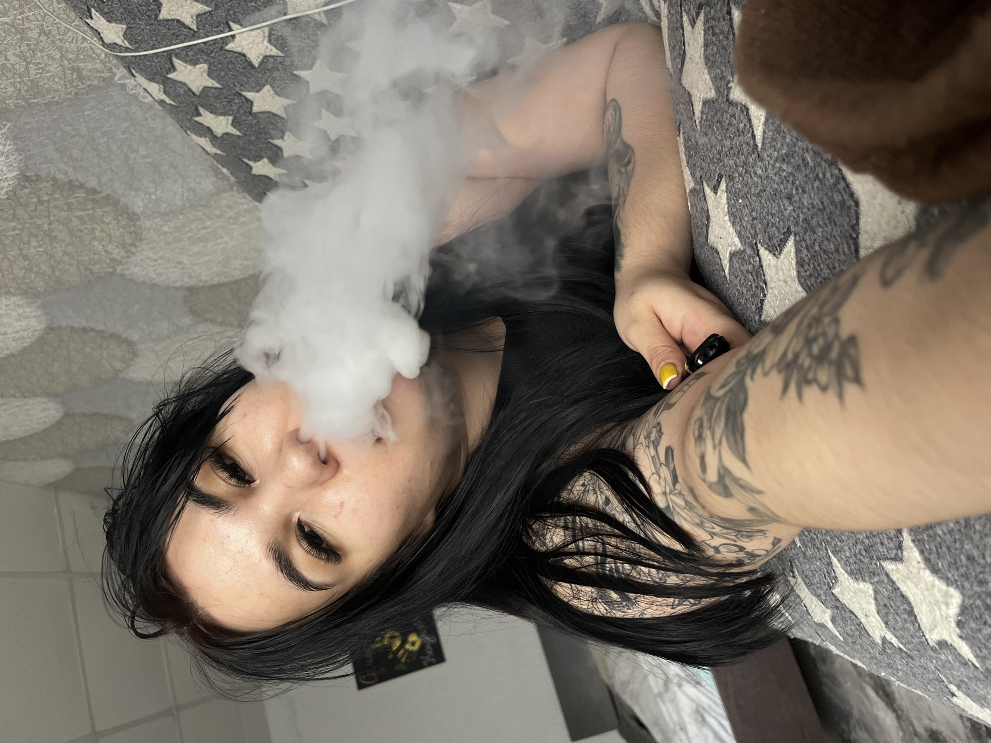 A woman with black hair and tattoos on her arms is lying on a gray blanket with white stars. She is exhaling a large plume of white vapor. She is holding a black object, possibly a vape pen, in her right hand, with a yellow painted fingernail visible. A picture is visible in the background. The scene appears to be indoors. The city of Chaikovsky, Russia, is not discernible from the image.