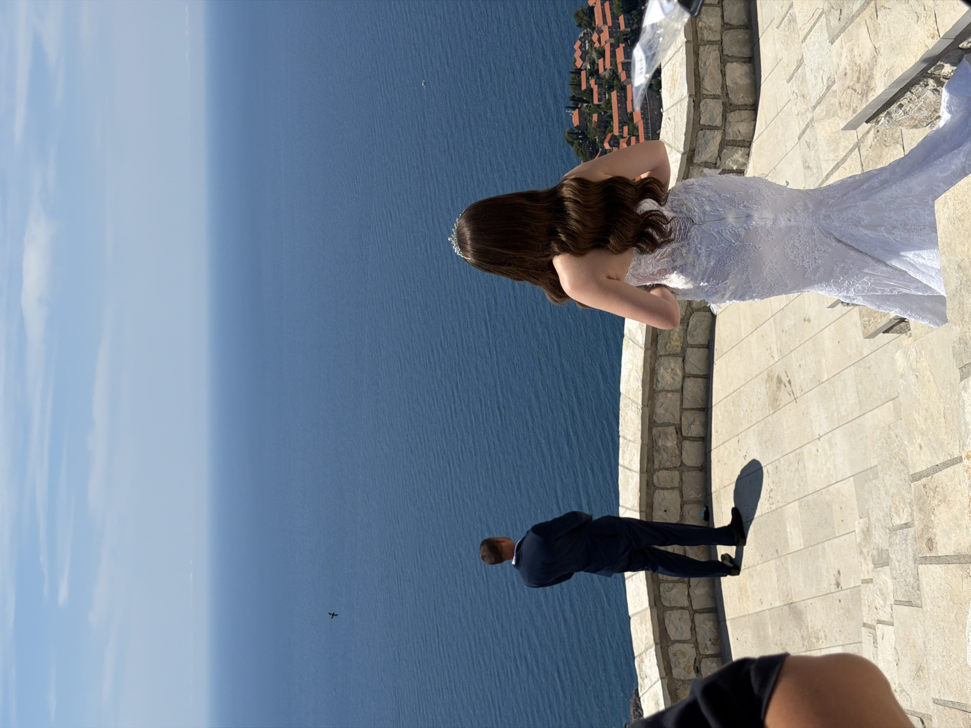 In Sveti Stefan, Montenegro, a bride in a white lace wedding dress and a groom in a dark suit stand on stone steps overlooking the Adriatic Sea. The bride, viewed from behind, has long, wavy brown hair and a tiara. The groom, also viewed from behind, stands with his arms crossed. A bird flies in the distance above the calm, blue water. In the background, the iconic red-roofed buildings of Sveti Stefan are visible on a peninsula. The scene is bathed in bright sunlight under a clear blue sky with sparse clouds.