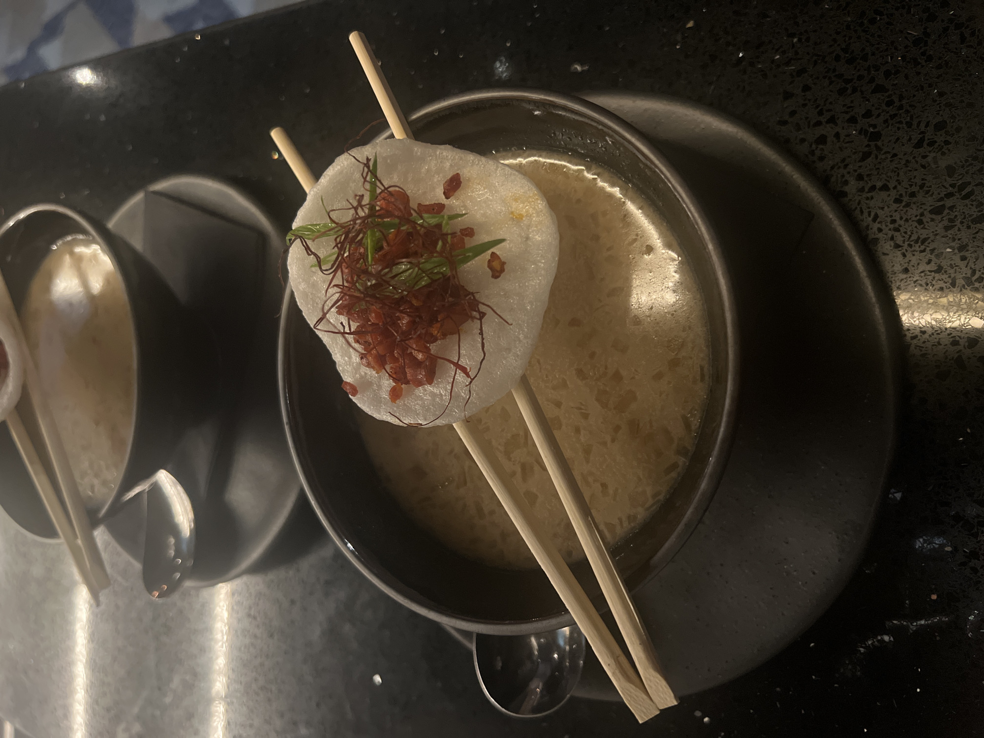 The image shows a close-up, high-angle shot of two small, black bowls containing soup. Both bowls are served on a dark, textured surface. The larger bowl features a white, circular topping pierced by two chopsticks. The topping is garnished with red and green herbs. A smaller bowl also contains soup and is served with chopsticks. The setting appears to be a restaurant, but the precise location in Sofia, Bulgaria, cannot be determined from the image. There are no people visible. The primary objects are the bowls, soup, chopsticks, and topping. The action involves serving the soup.