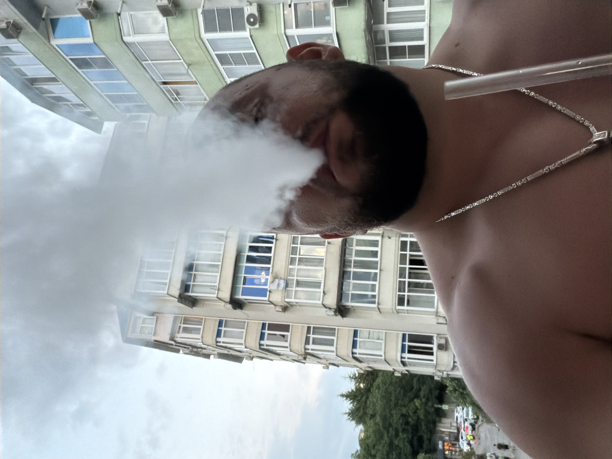A man with a beard is exhaling smoke, obscuring his face, while holding a hookah hose. He is shirtless and wearing a silver necklace. In the background, there are apartment buildings and trees. The setting appears to be a city, possibly Sochi, Russia, given the architectural style and greenery. The sky is overcast.