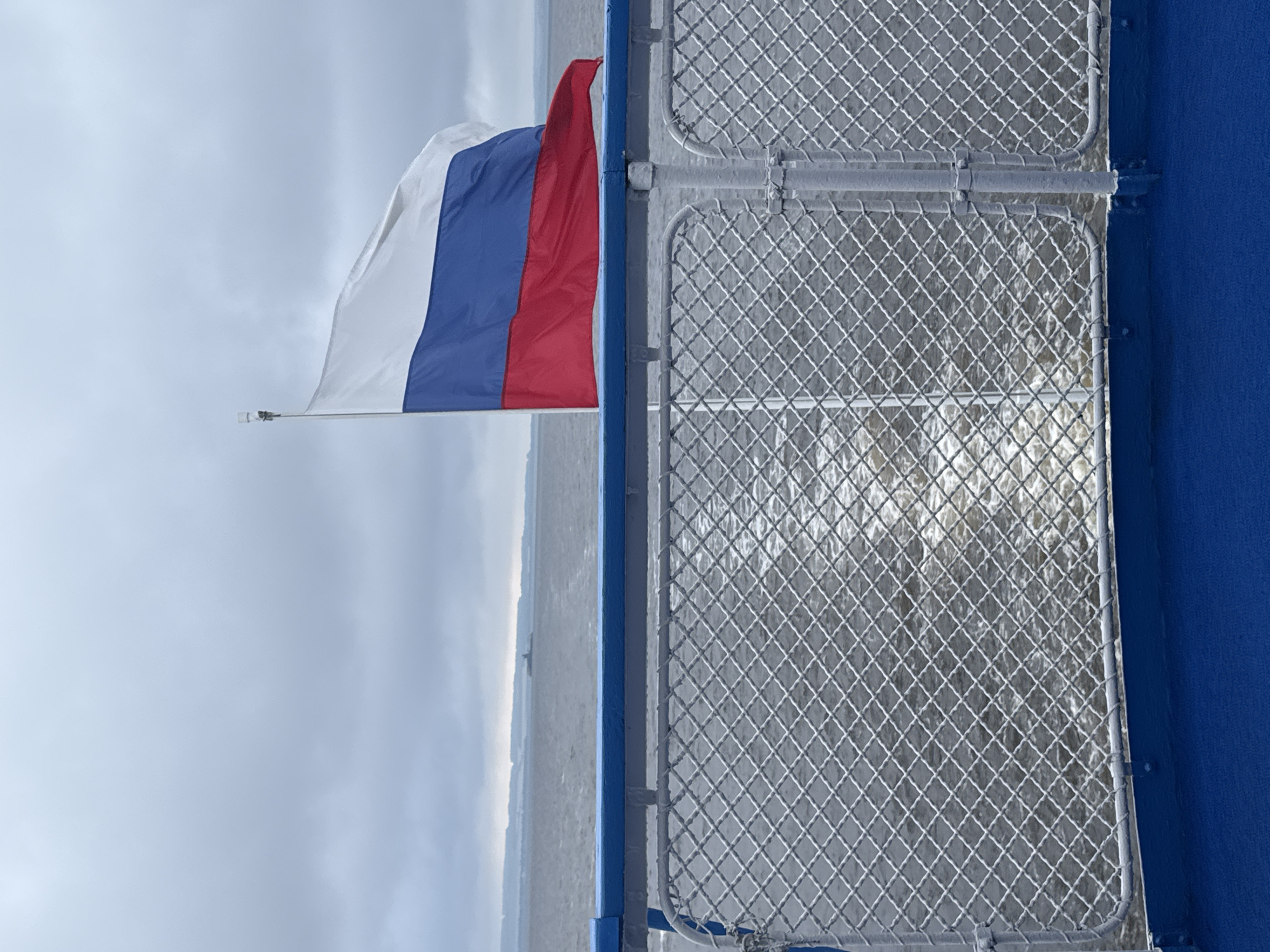 The image captures a view from the deck of a vessel, likely a ferry, moving through a body of water. In the foreground, a blue painted railing runs horizontally, topped by a wire mesh fence. The water behind the fence is churned into white foam, indicating the vessel's movement. In the background, a Russian flag, with its white, blue, and red horizontal stripes, flies from a flagpole. The flag is partially unfurled, catching the wind. Beyond the water, a hazy shoreline with a faint structure is visible under an overcast sky. The scene suggests a journey on a waterway near Laishevo, Russia, on a cloudy day.