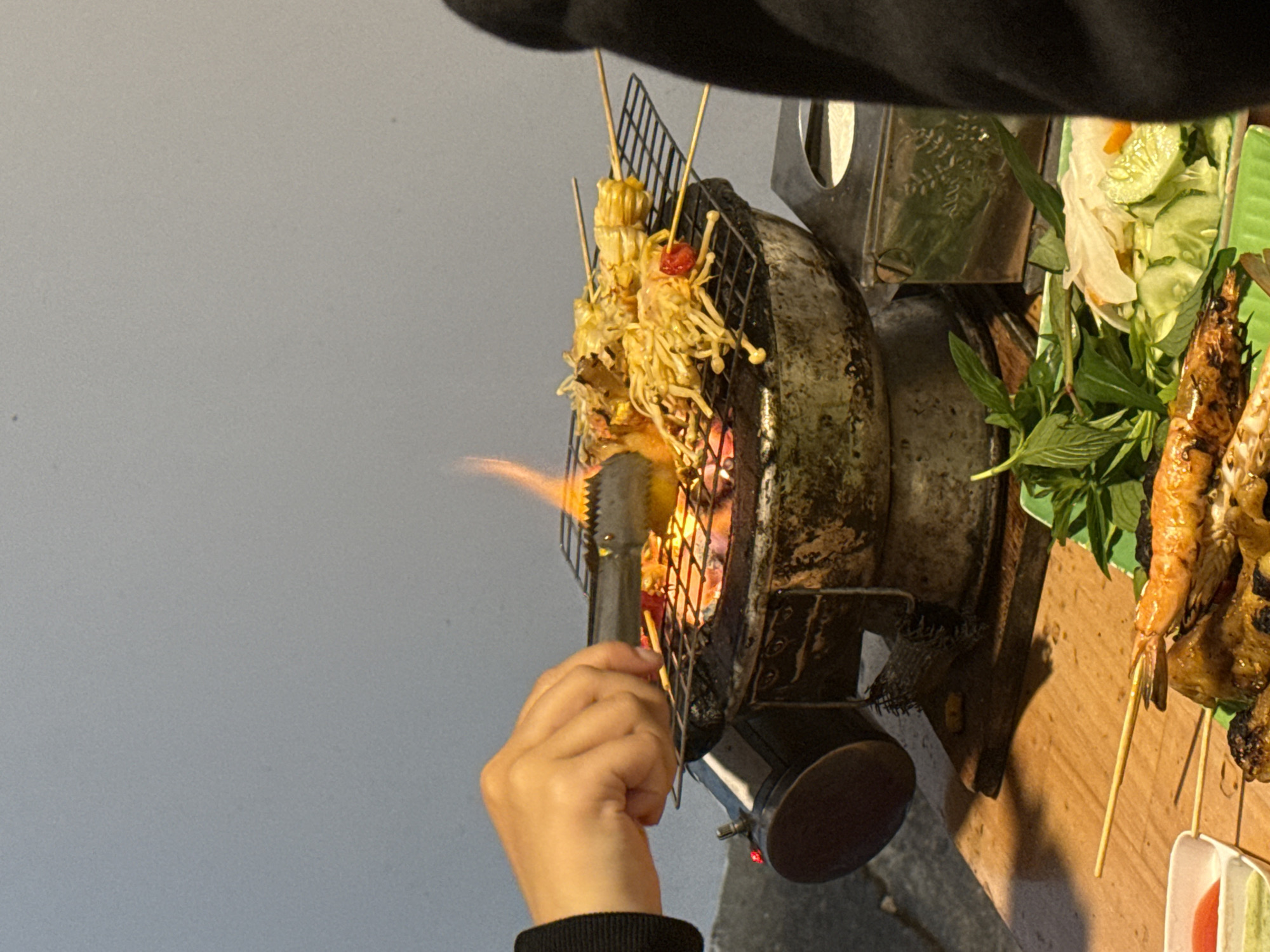 A person’s hand in a black sweater sleeve is adjusting food on skewers over a small grill with visible flames, in Da Lat, Vietnam. The food includes skewers of mushrooms and corn. Adjacent to the grill is a plate of shrimp skewers with fresh basil, lettuce, and sliced cucumbers. The background is a solid light grey wall.