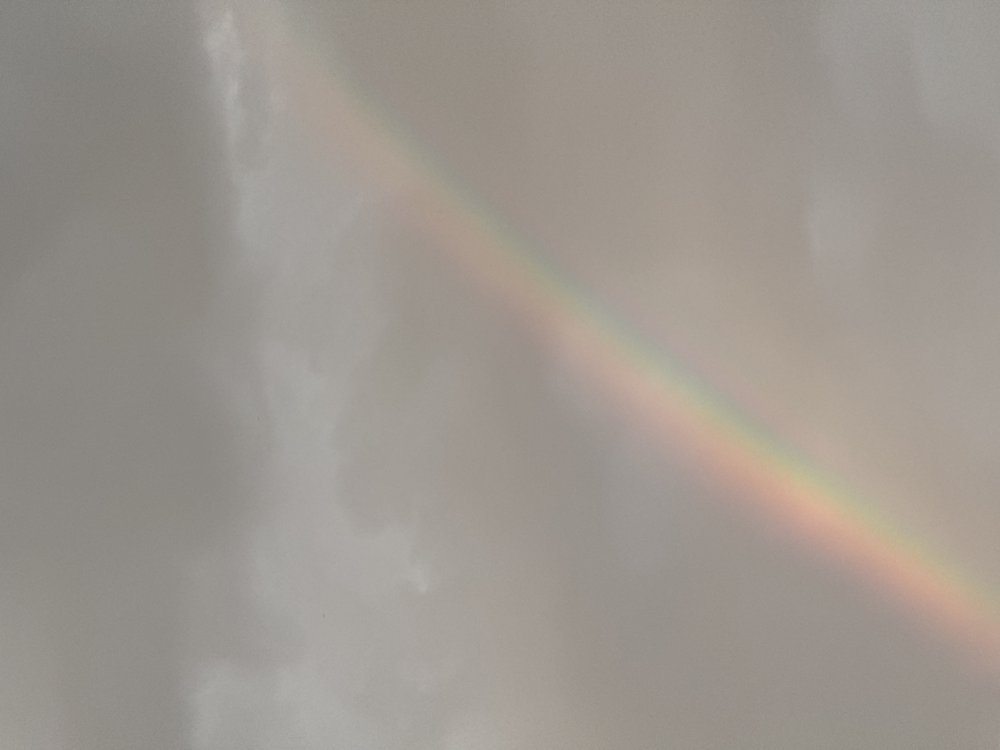 A faint rainbow arcs across a hazy, overcast sky. The clouds are a muted gray, suggesting a recent or impending rain. The rainbow's colors are soft and diffused, appearing as bands of pastel red, orange, yellow, green, and possibly hints of blue. There are no people or discernible structures visible in the image, the scene is solely focused on the atmospheric phenomenon. The location is not specified in the image.