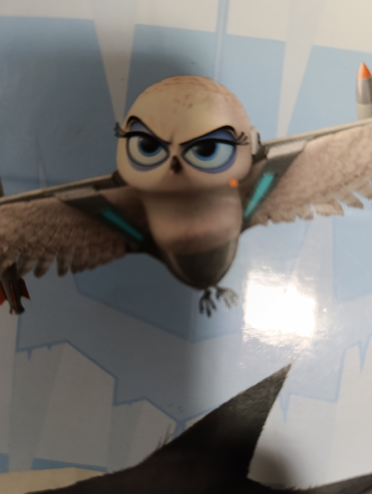 A close-up shot depicts a cartoon owl-like figure with airplane wings. The figure has large, blue eyes with prominent eyelashes and an angry expression. Its body is brown and grey, and the wings extend outward with grey and black accents and a teal stripe. An orange and grey rocket-like object is attached to the end of one wing. In the background, there is a stylized depiction of a city with white and blue buildings. A dark triangular shape is visible in the lower foreground.