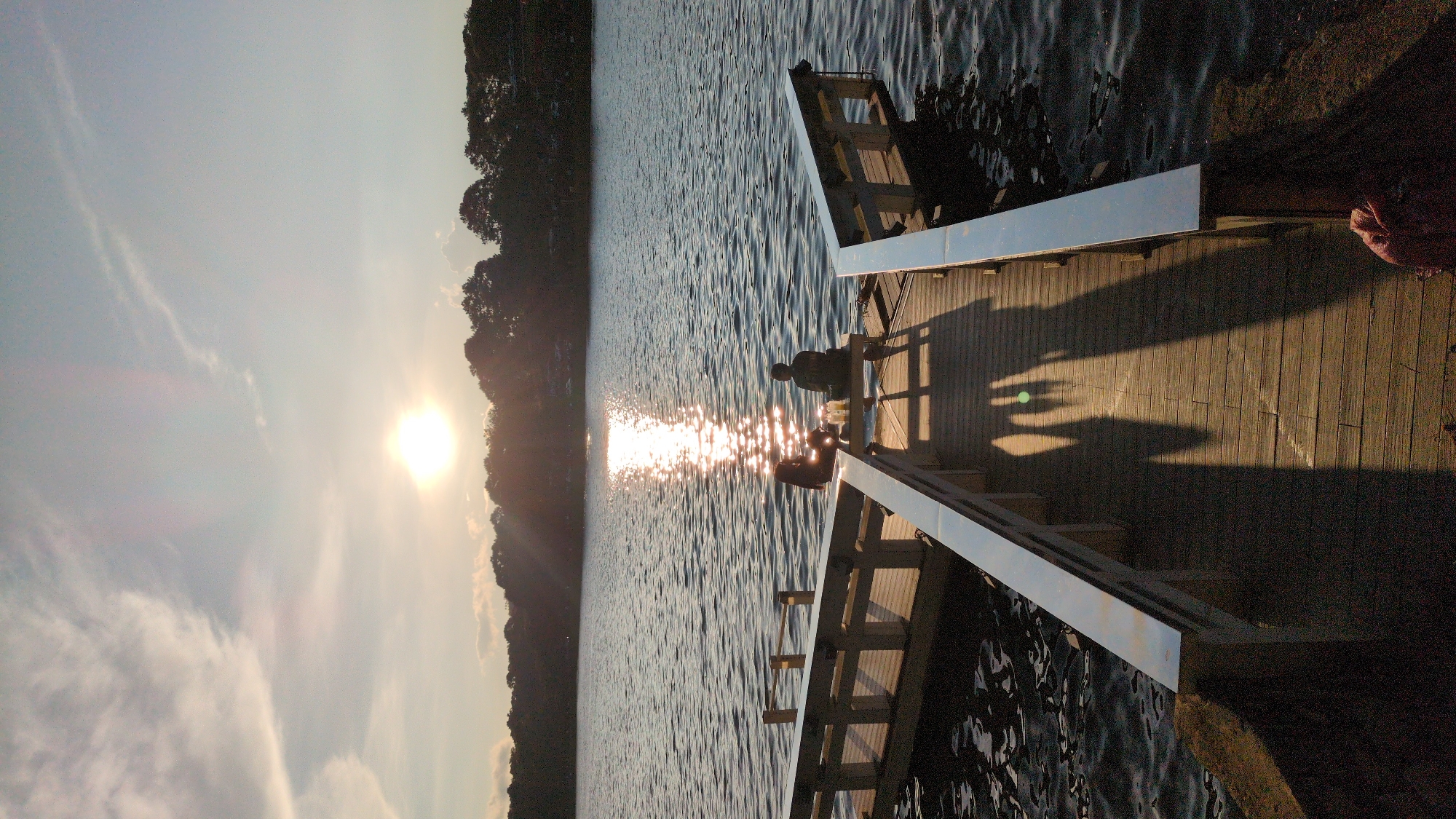 The image shows a wooden dock extending into a body of water during sunset. Two individuals are seated on the dock, facing the water and the setting sun. The sun is visible in the sky, casting a bright reflection on the water's surface. Trees line the distant shore. The dock has railings and a wooden plank surface. The lighting suggests late afternoon or early evening. There is no indication that this location is in Da Lat, Vietnam; it appears to be a generic lake or coastal scene.