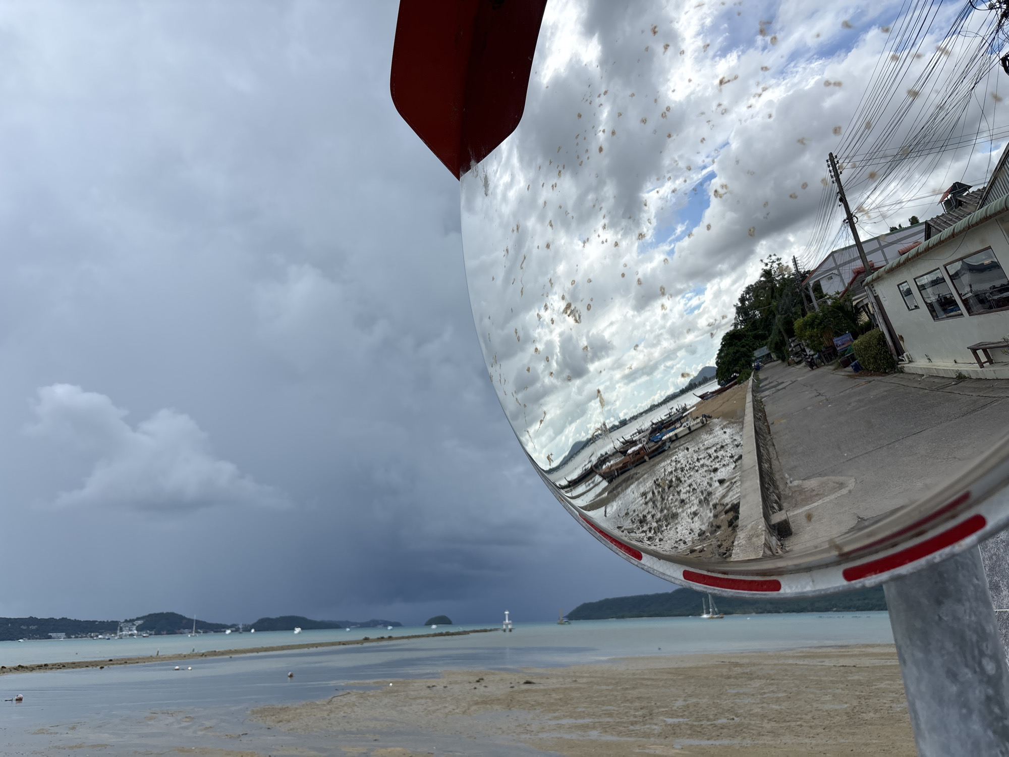 An exterior, eye-level, wide shot captures a convex mirror reflecting a street scene in Rawai, Thailand, on a cloudy day. The mirror is positioned on the right side of the frame, tilted forward and slightly to the left, obscuring the main view of the street which curves away to the right. The reflection shows a paved road with buildings on the right side. Several power lines are strung between poles along the road, and parked motorcycles are visible. A white building with large windows and a small bench outside is prominent. The left side of the mirror's reflection shows a shore with many boats docked and a wall along the water's edge. Beyond the boats, a body of water with several smaller boats and a distant lighthouse is visible. The sky in the reflection is partly cloudy with patches of blue. The foreground of the actual scene shows a muddy, sandy shore with shallow water. The sky in the upper left of the frame is overcast with dark clouds, suggesting impending rain. The mirror itself has a red border and several circular red reflectors along its lower rim. The mirror is attached to a gray pole.
