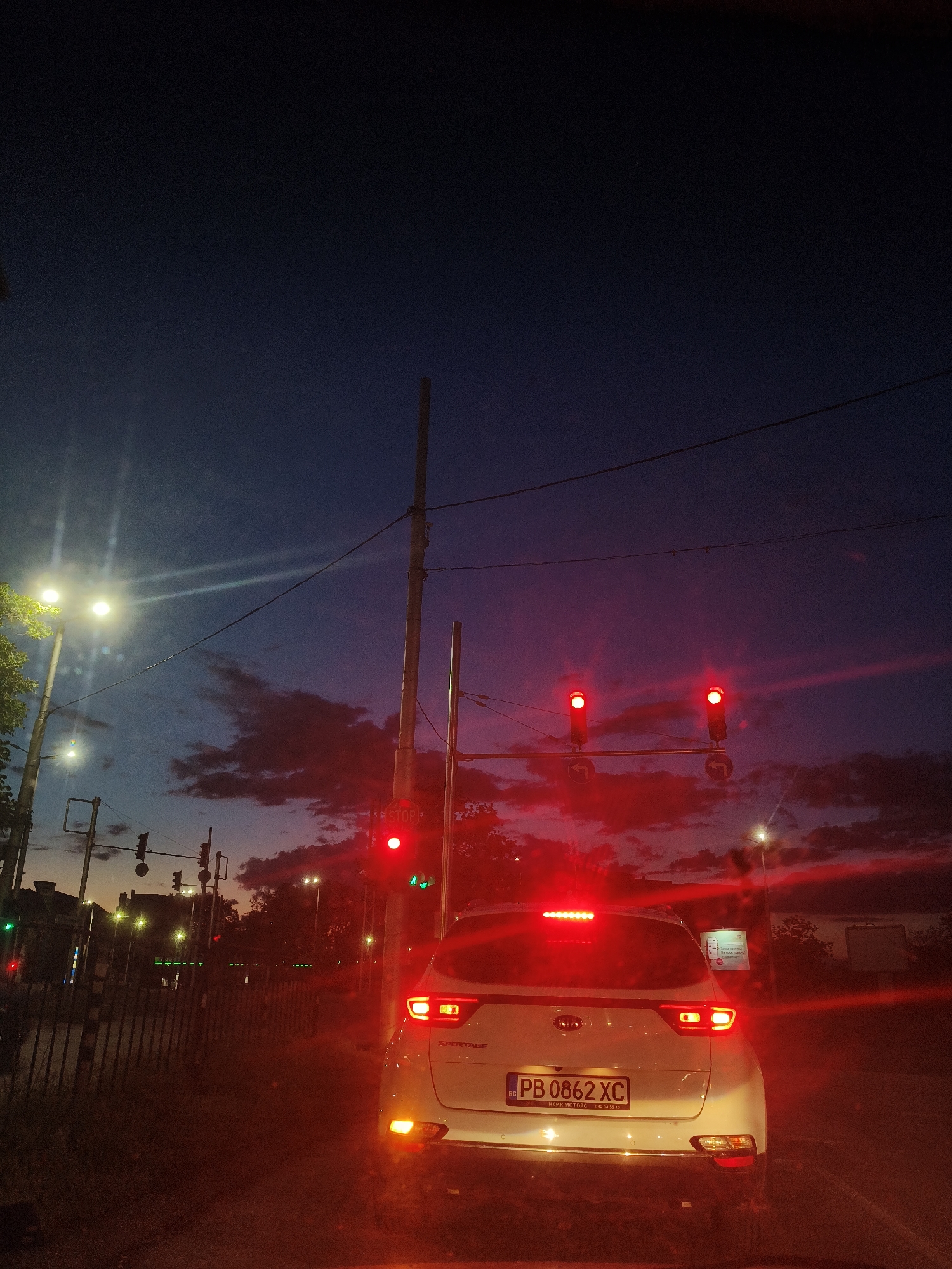 The image shows the rear of a white Kia Sportage with Bulgarian license plate "PB 0862 XC" at night. The car is stopped at a traffic light showing red. Other traffic lights are visible in the background. The location appears to be a street in Plovdiv, Bulgaria, given the license plate. The sky is dark with some clouds visible. There are power lines and poles present. There is a fence visible to the left of the car. The scene is illuminated by streetlights and the car's brake lights.