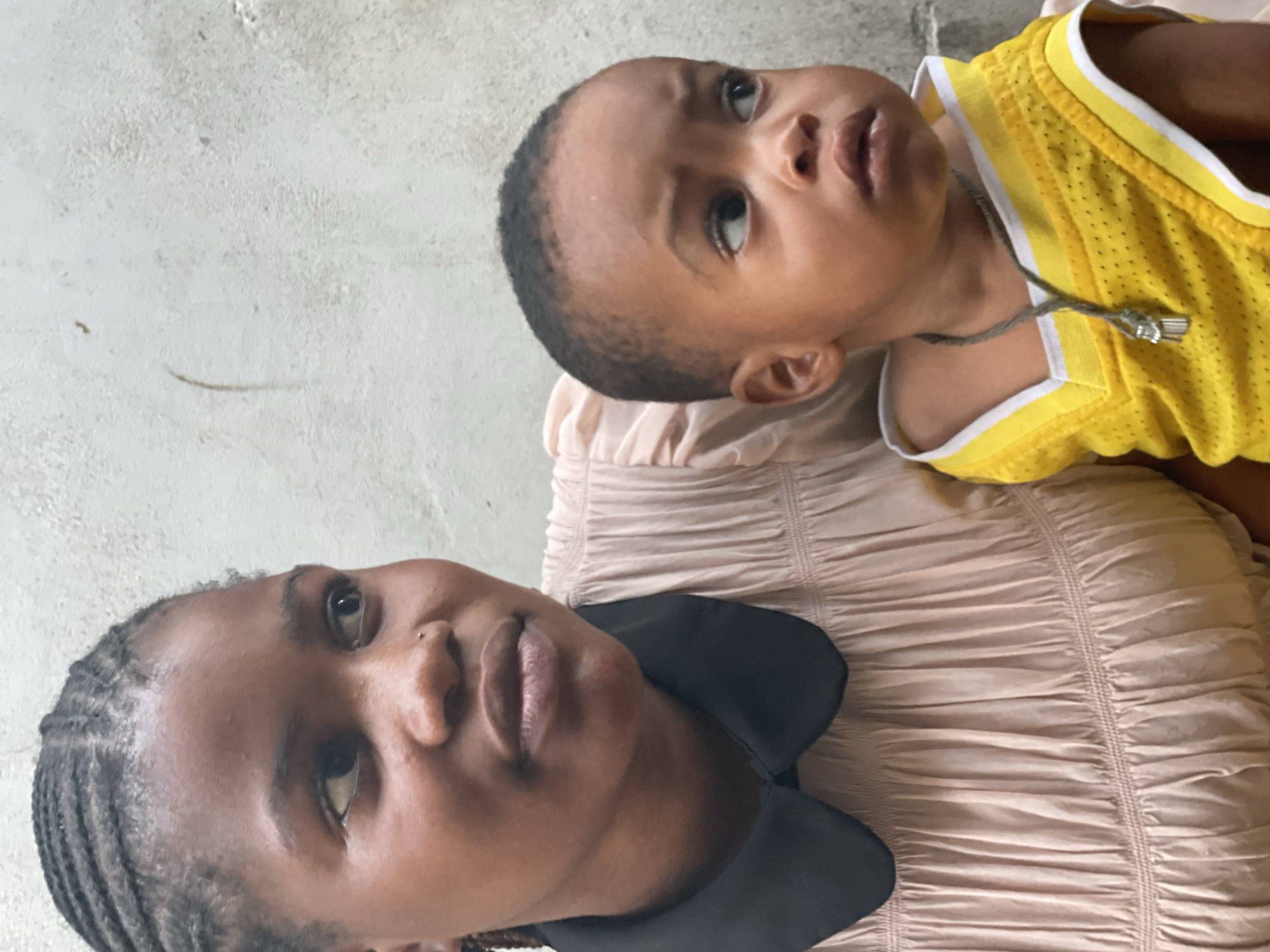 A medium shot depicts an adult woman and an infant. The woman, positioned on the left side of the frame, has dark skin, braided hair, and a nose piercing. She wears a peach-colored garment with a black collar. The infant, situated above the woman, has a light brown complexion and wears a yellow and white tank top with a necklace. The background appears to be a wall of cement. The location is presumed to be Badagry, Nigeria.